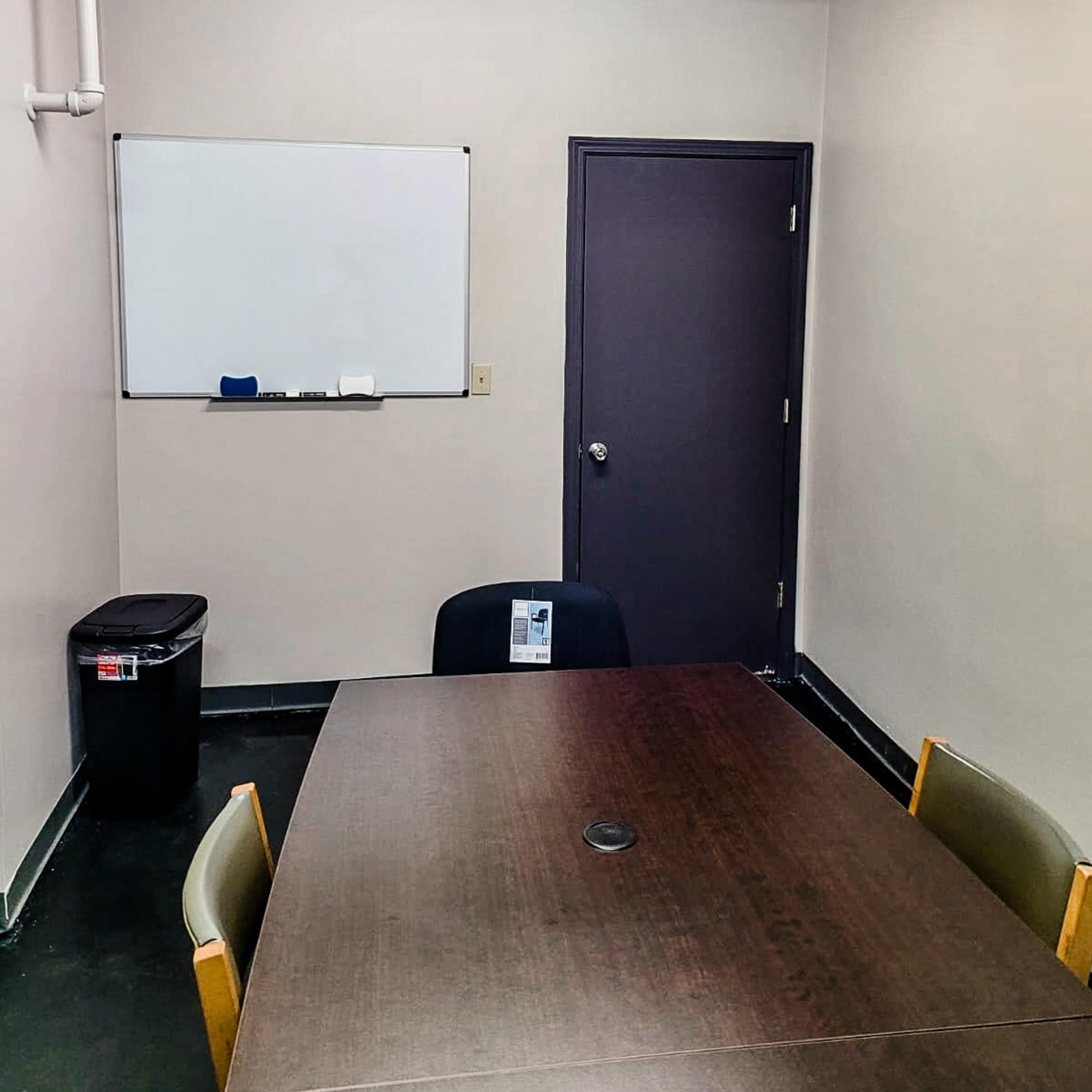 A small meeting room with a round table, four chairs, a whiteboard on the wall, a trash can, and a door.