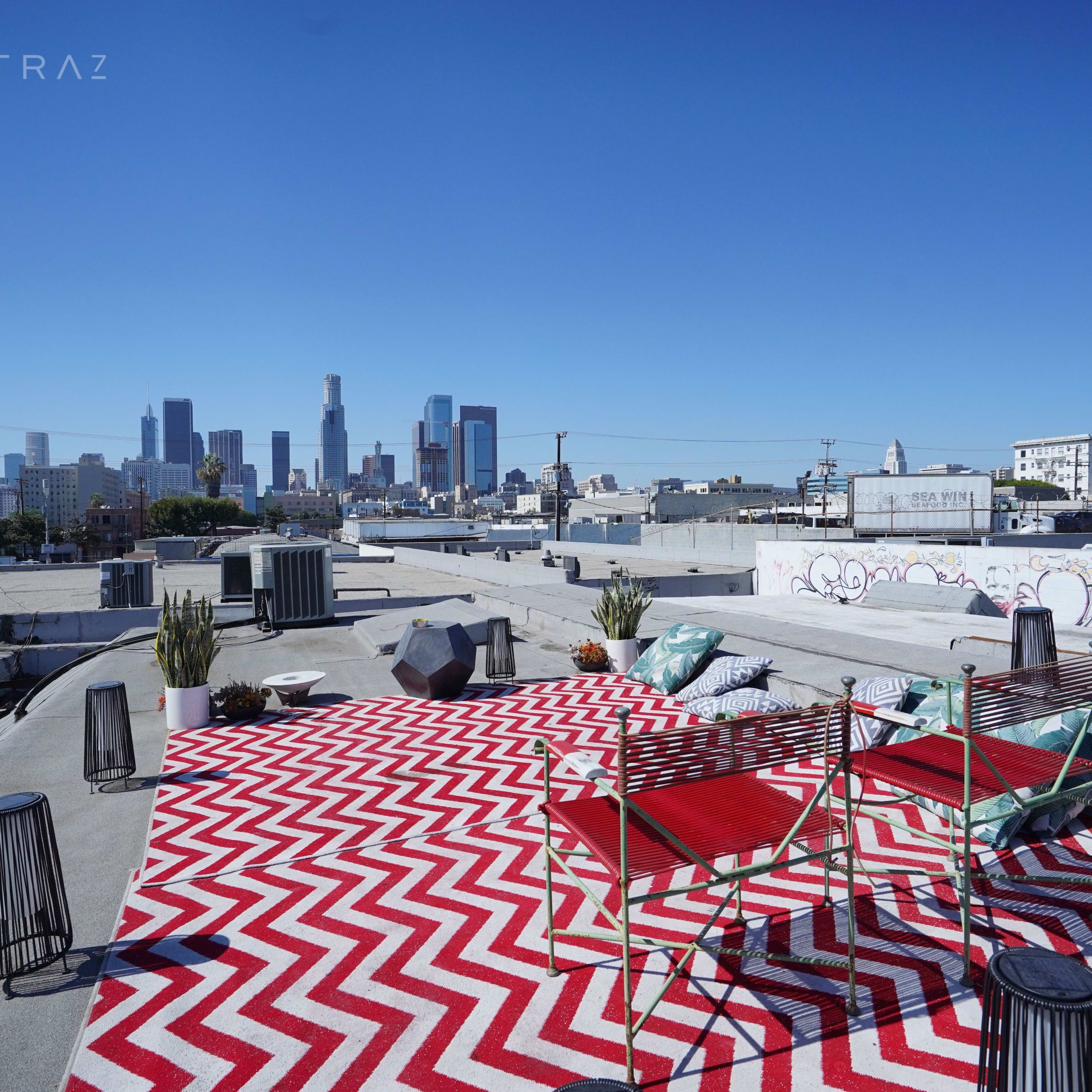 A rooftop terrace with a red and white zigzag rug, two chairs, plants, and a view of the Los Angeles skyline.