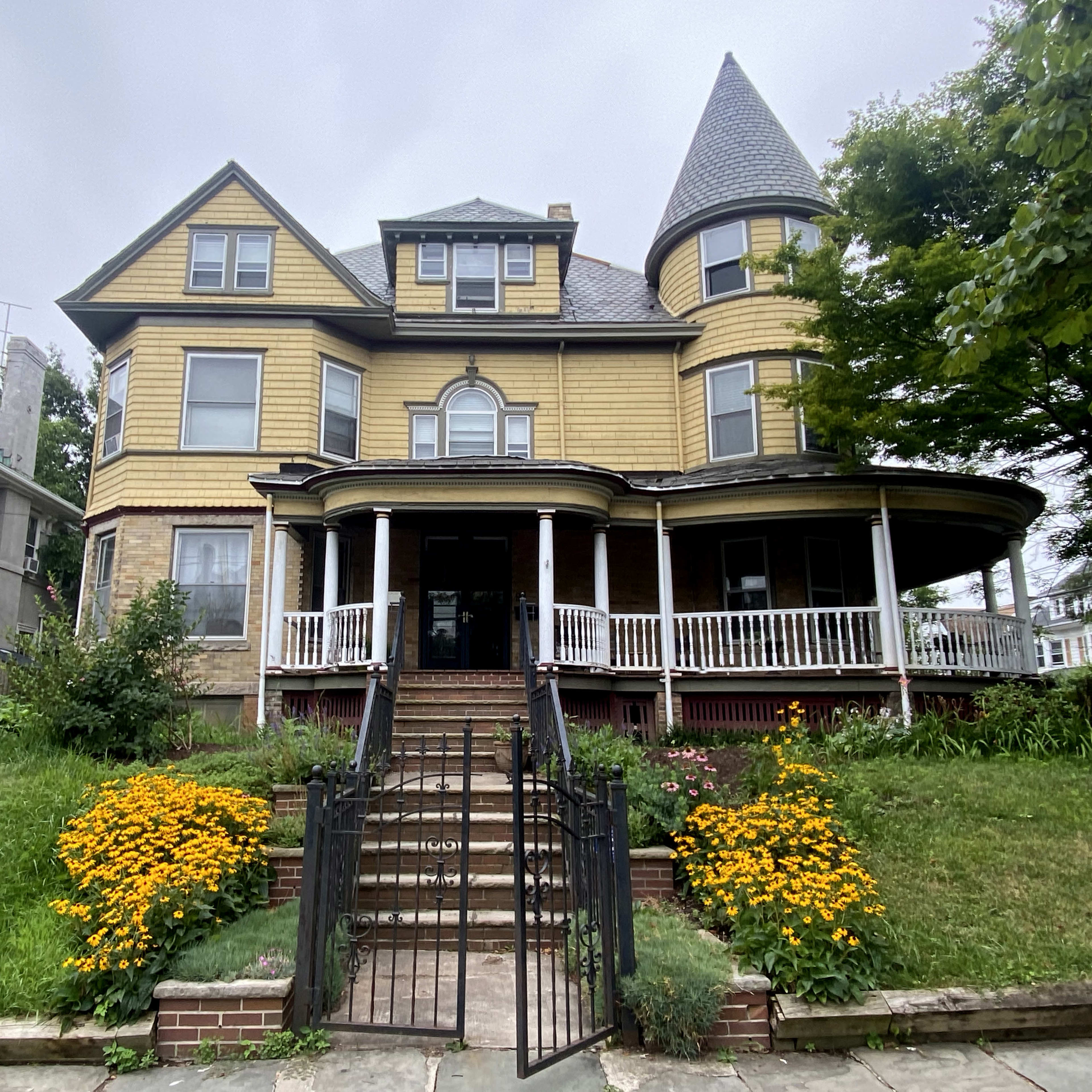 A large, multi-story Victorian house with a conical tower and a wraparound porch is surrounded by a garden featuring bright yellow flowers.