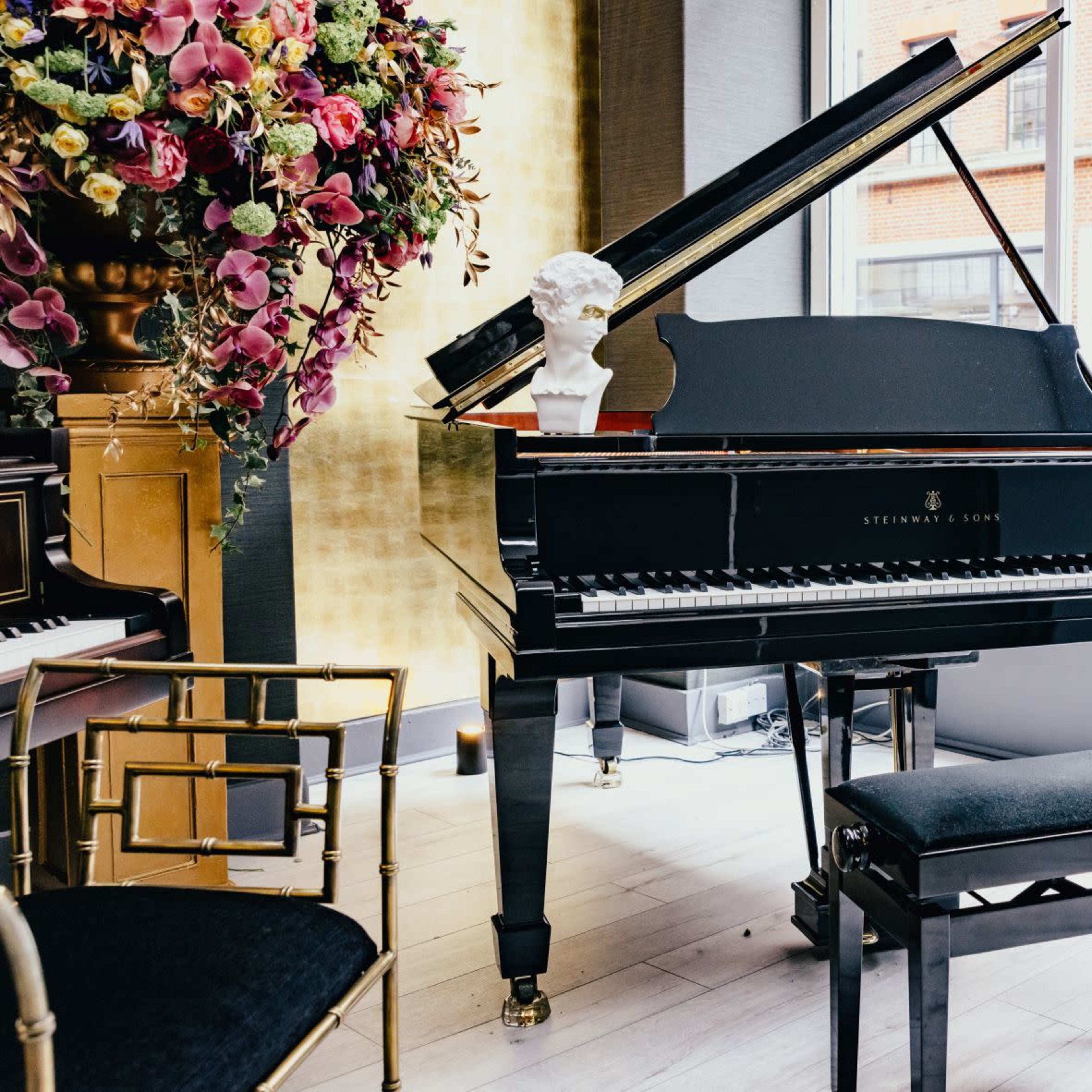 A black grand piano sits beside a large window, adorned with a floral arrangement and a decorative bust.