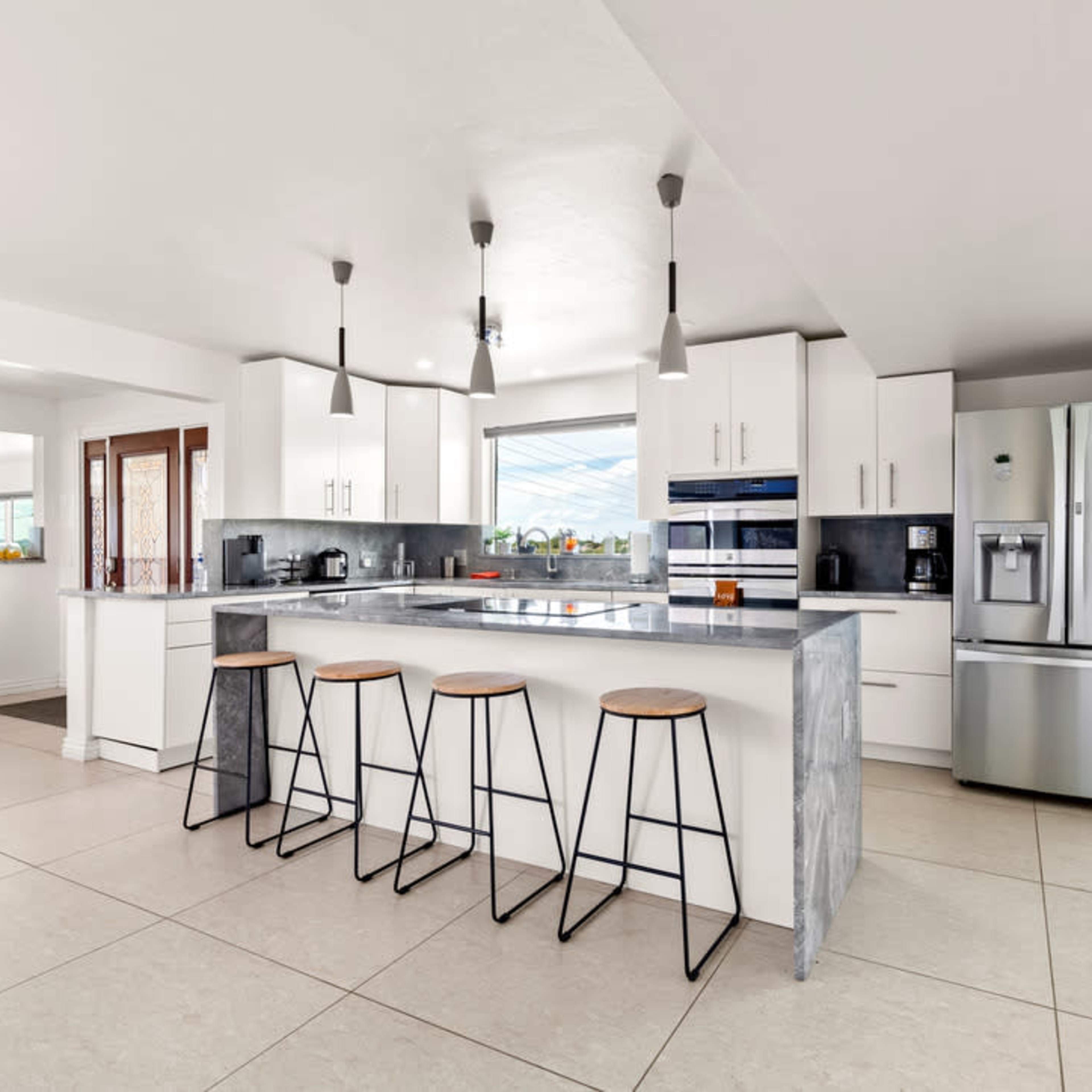A modern kitchen features a central island with three stools, white cabinetry, stainless steel appliances, and large windows allowing natural light.
