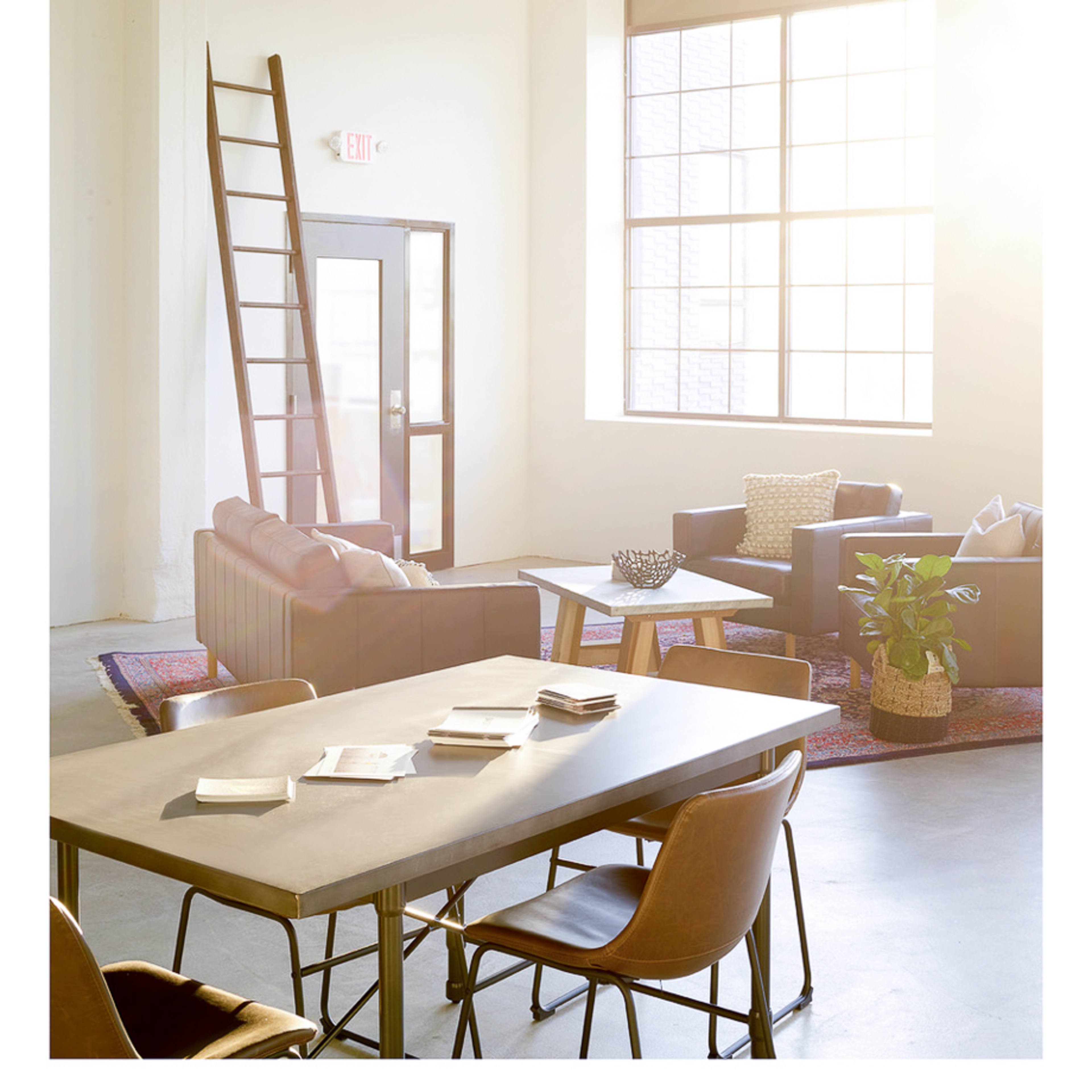 A bright, modern living space features a table with chairs, a sofa, and large windows letting in sunlight.