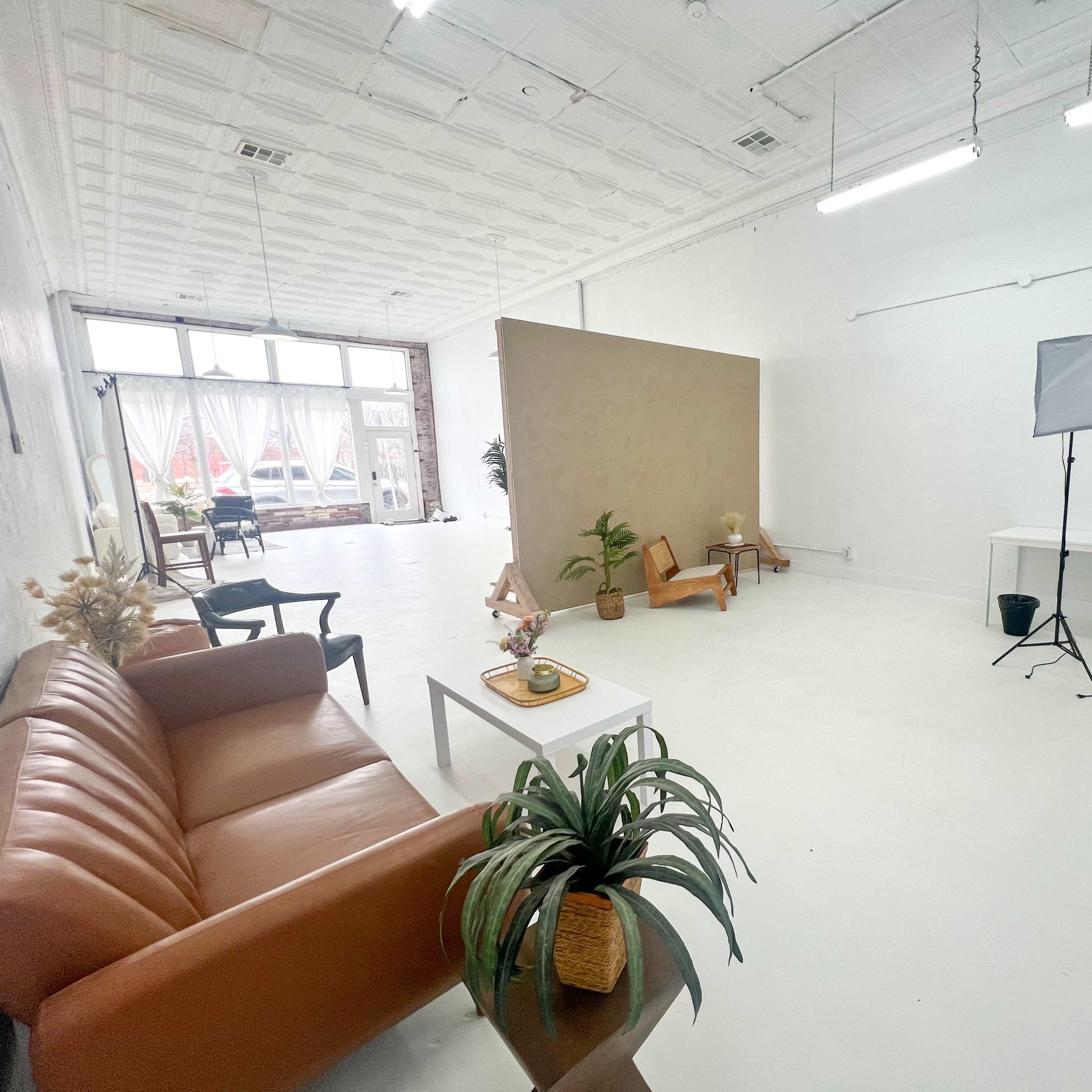 A bright, minimalist studio space featuring a brown sofa, two chairs, a photography light setup, and a neutral backdrop.