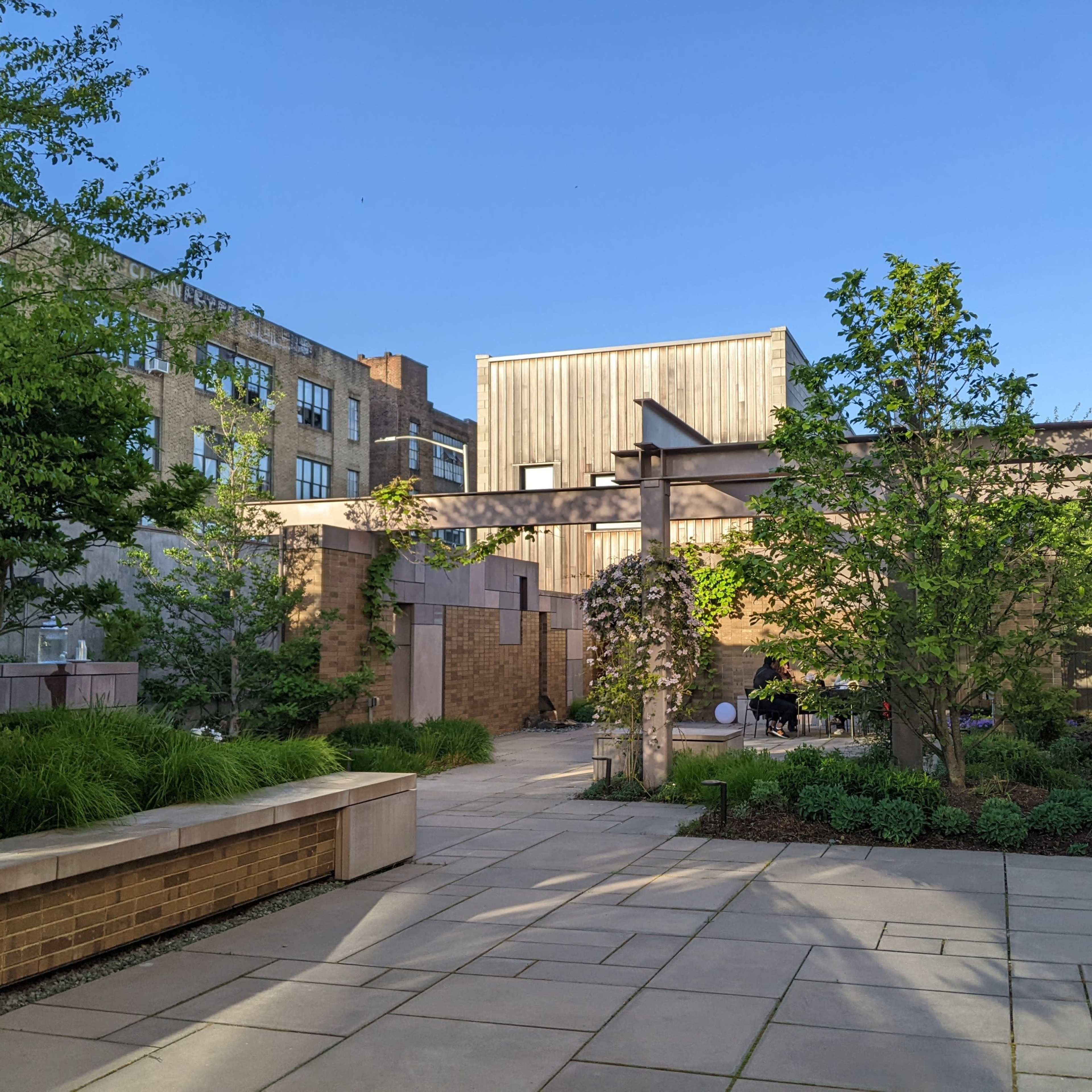 A landscaped urban courtyard with stone pathways, greenery, and modern architectural elements, surrounded by brick and wooden buildings.