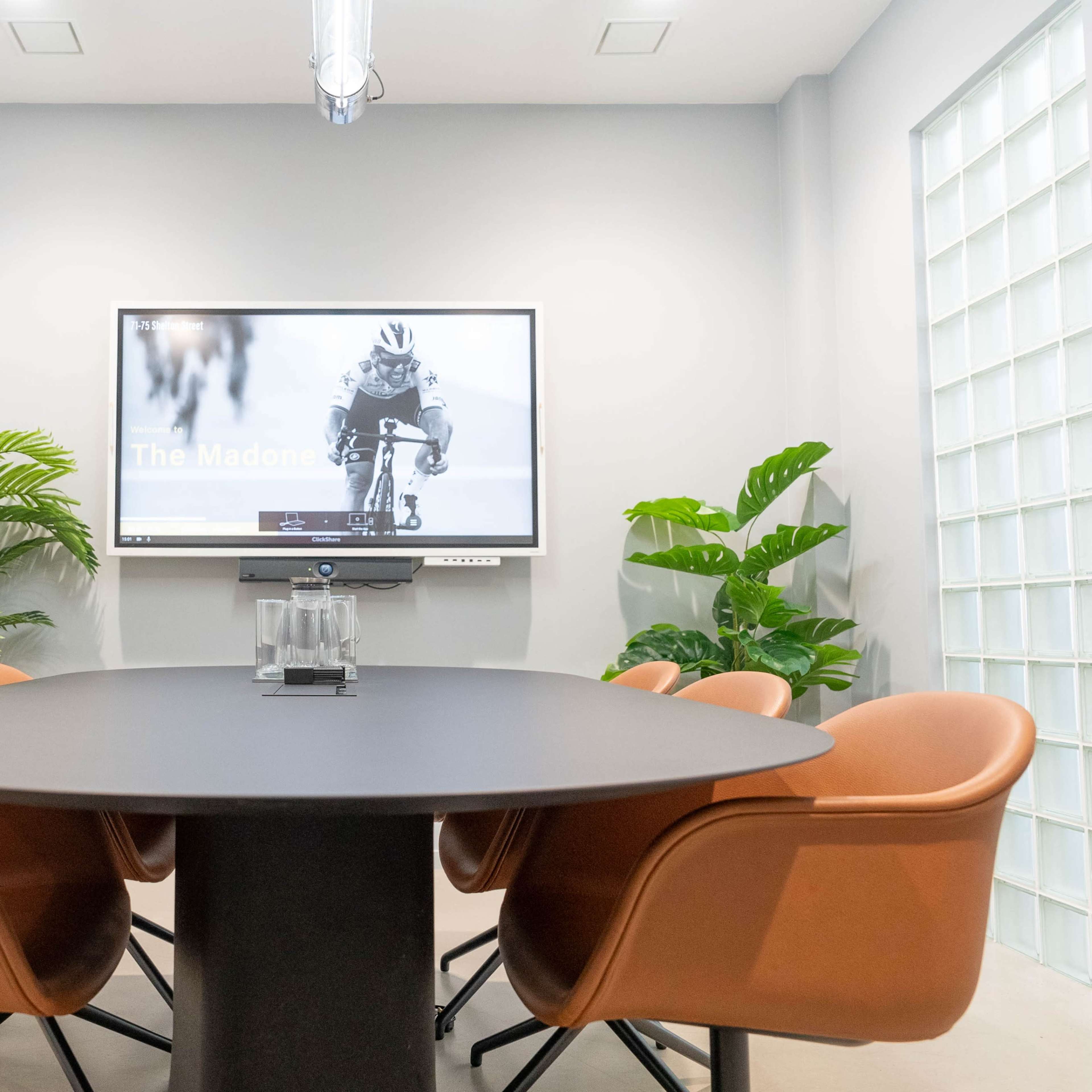 A conference room features a round table surrounded by brown chairs, with a wall-mounted screen displaying an image of a cyclist and potted plants in the background.