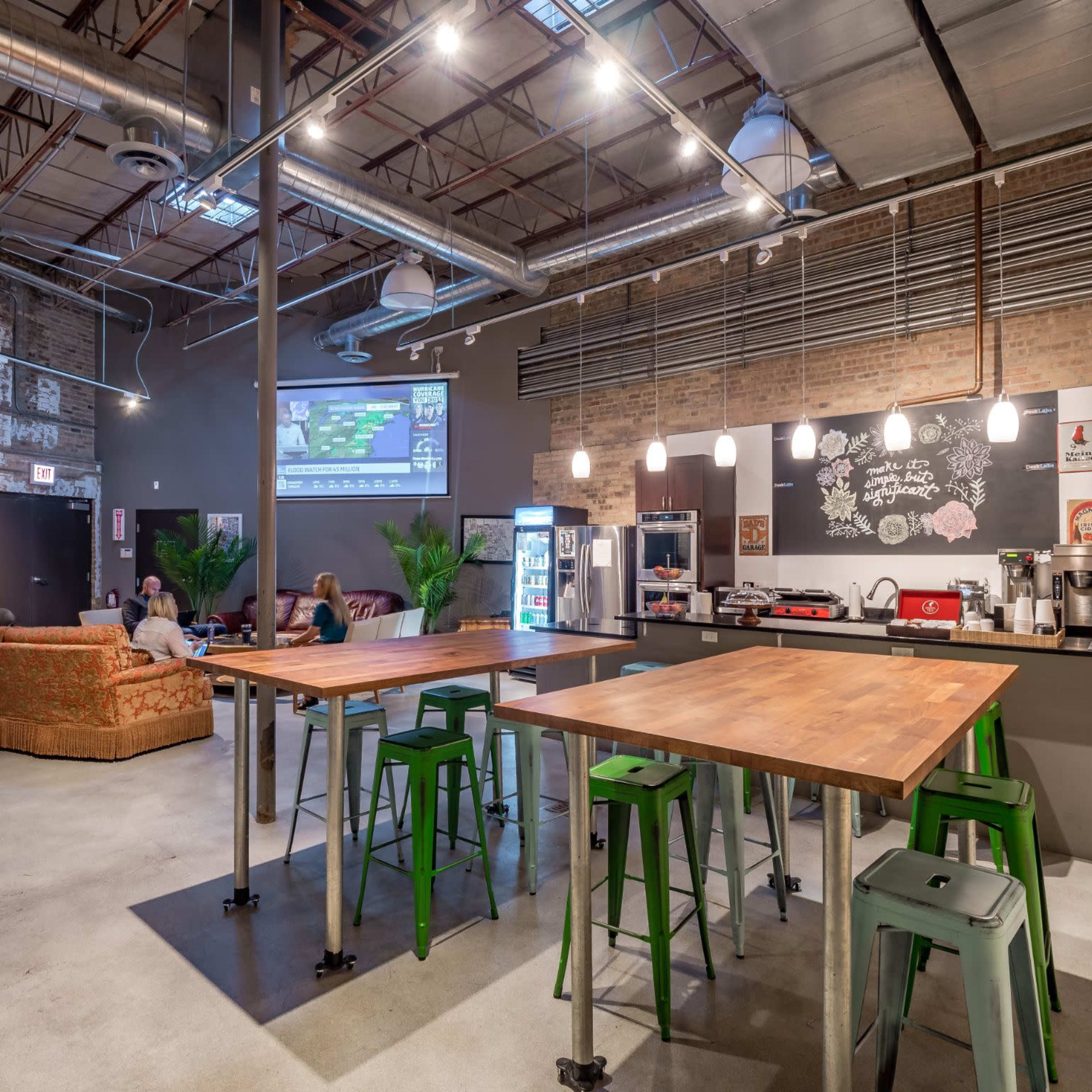 A spacious cafe features industrial decor with high tables, metal stools, a coffee machine, and a large screen displaying content on a brick wall.