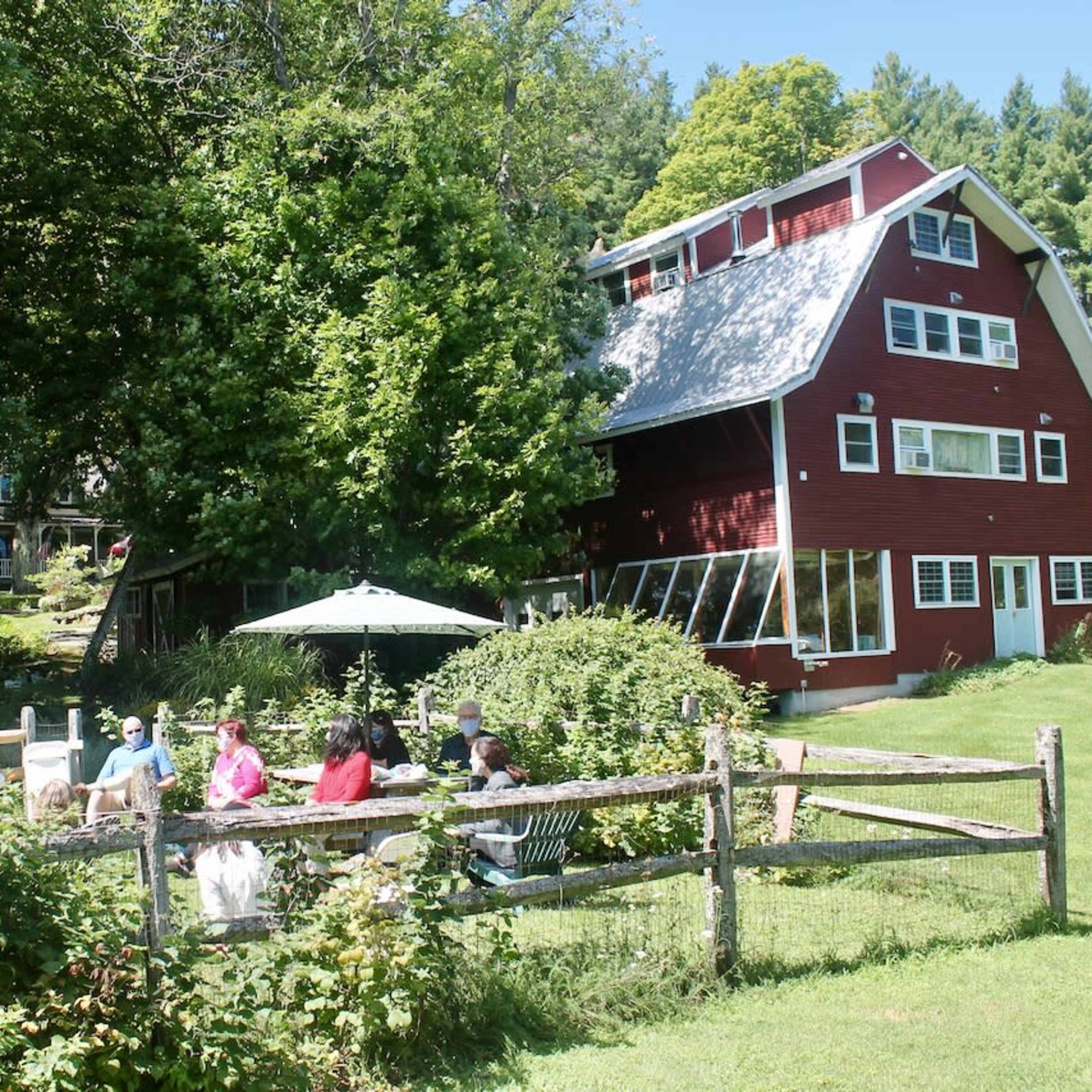 A group of people sits around a table under an umbrella near a large red house surrounded by trees and greenery.