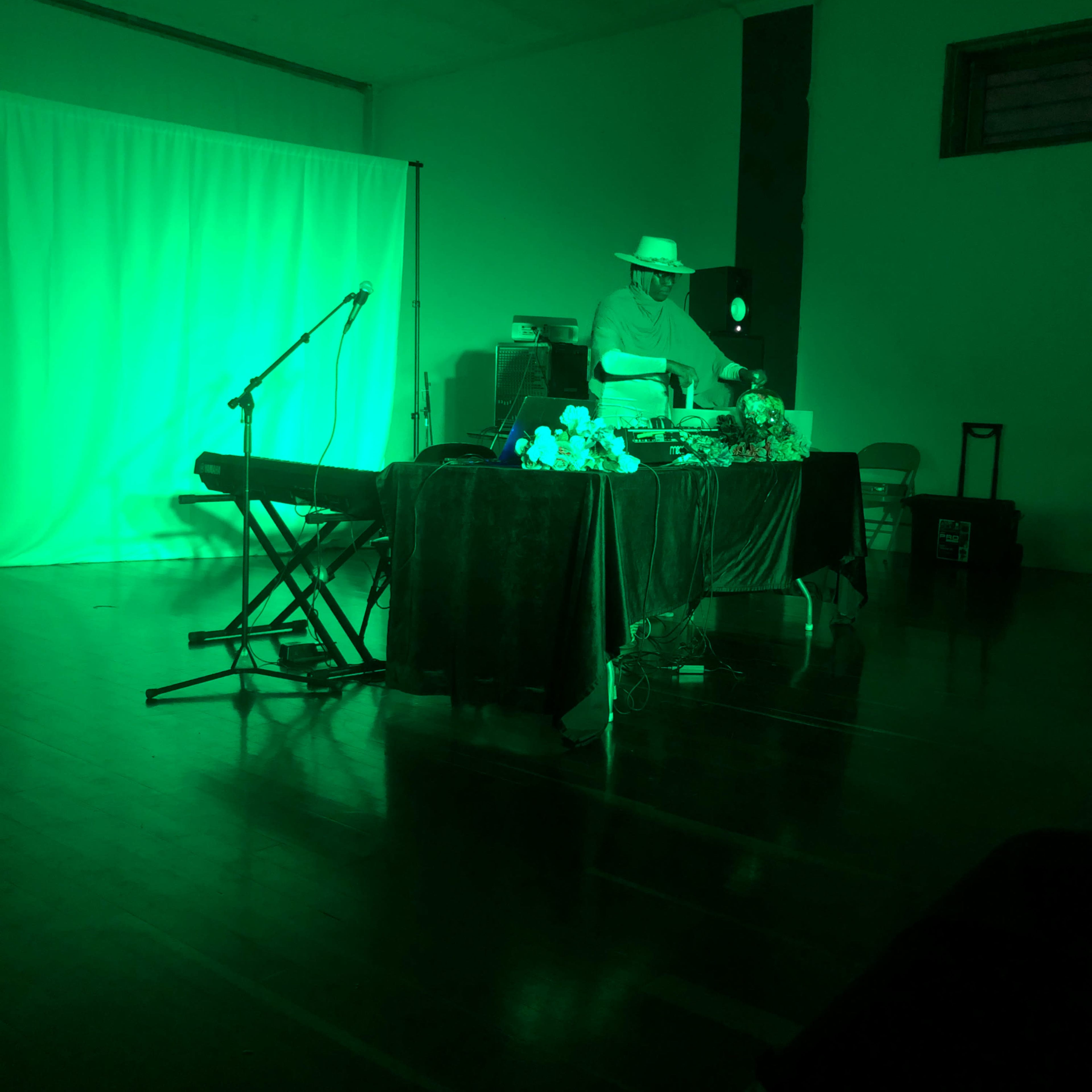 A person is performing behind a table covered with flowers and equipment under green lighting in a room with a white backdrop.
