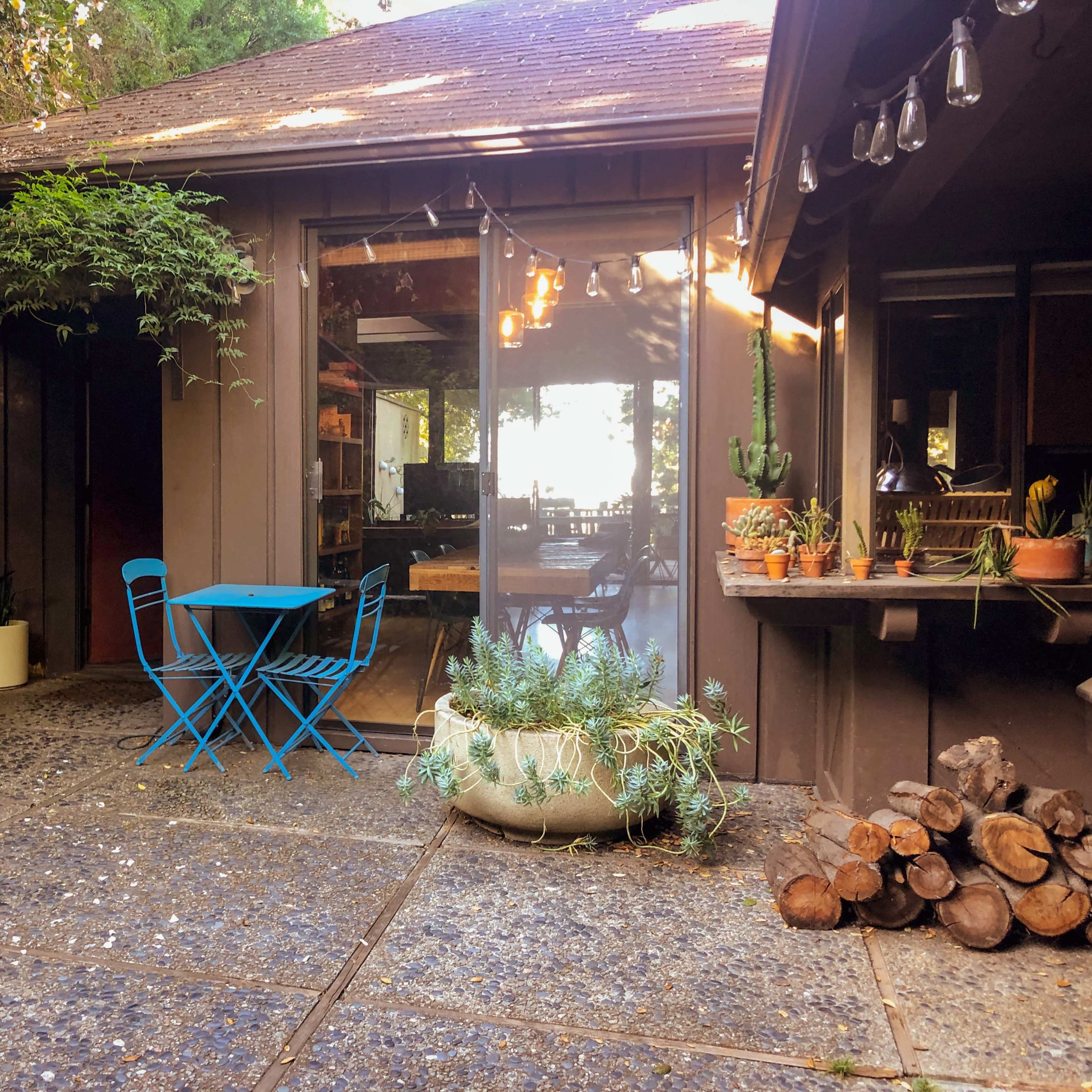 A small outdoor patio features a blue table and chairs, potted plants, a stack of firewood, and a dog standing nearby.