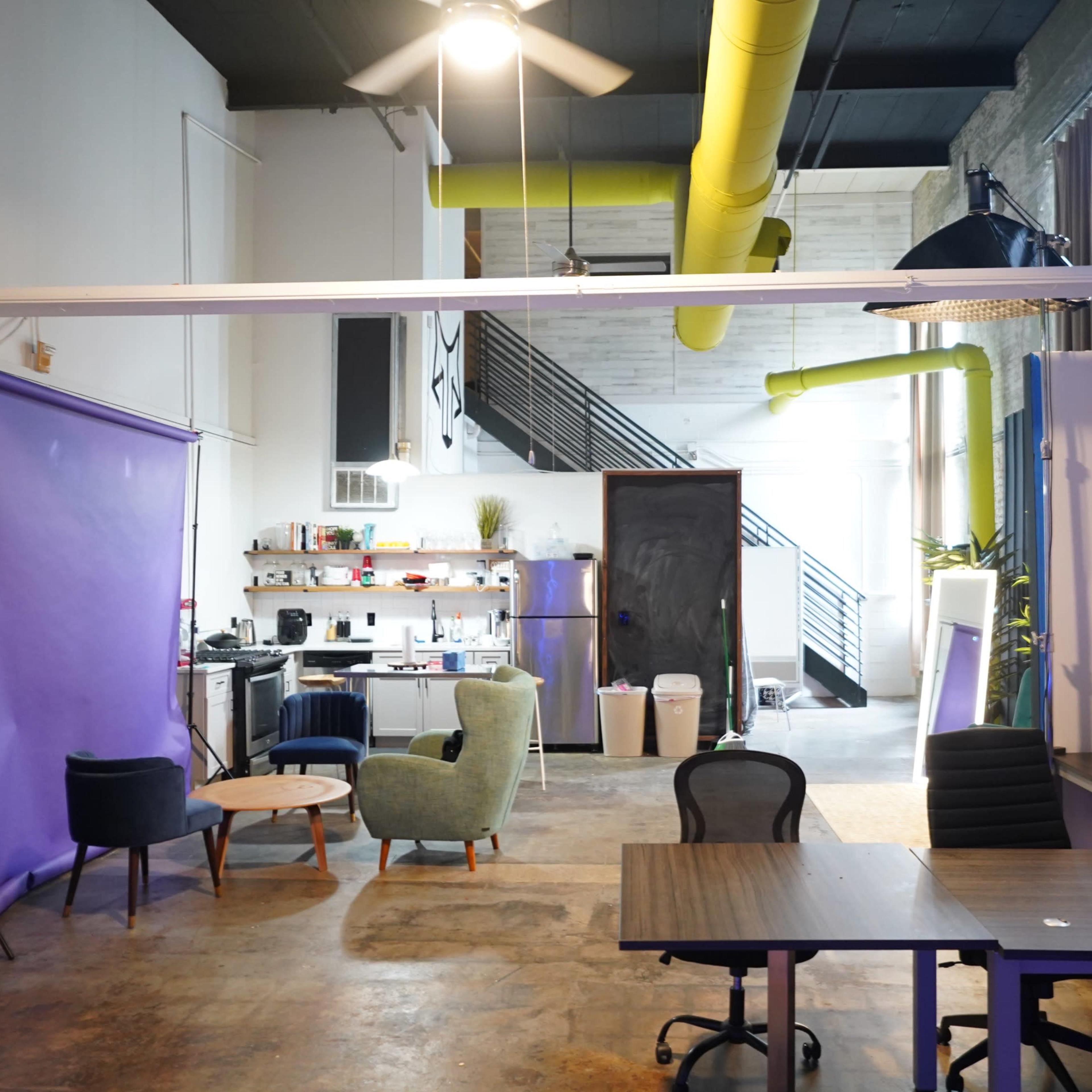 A modern office space features seating areas, a kitchen, and a staircase, with yellow ductwork adding color to the industrial aesthetic.
