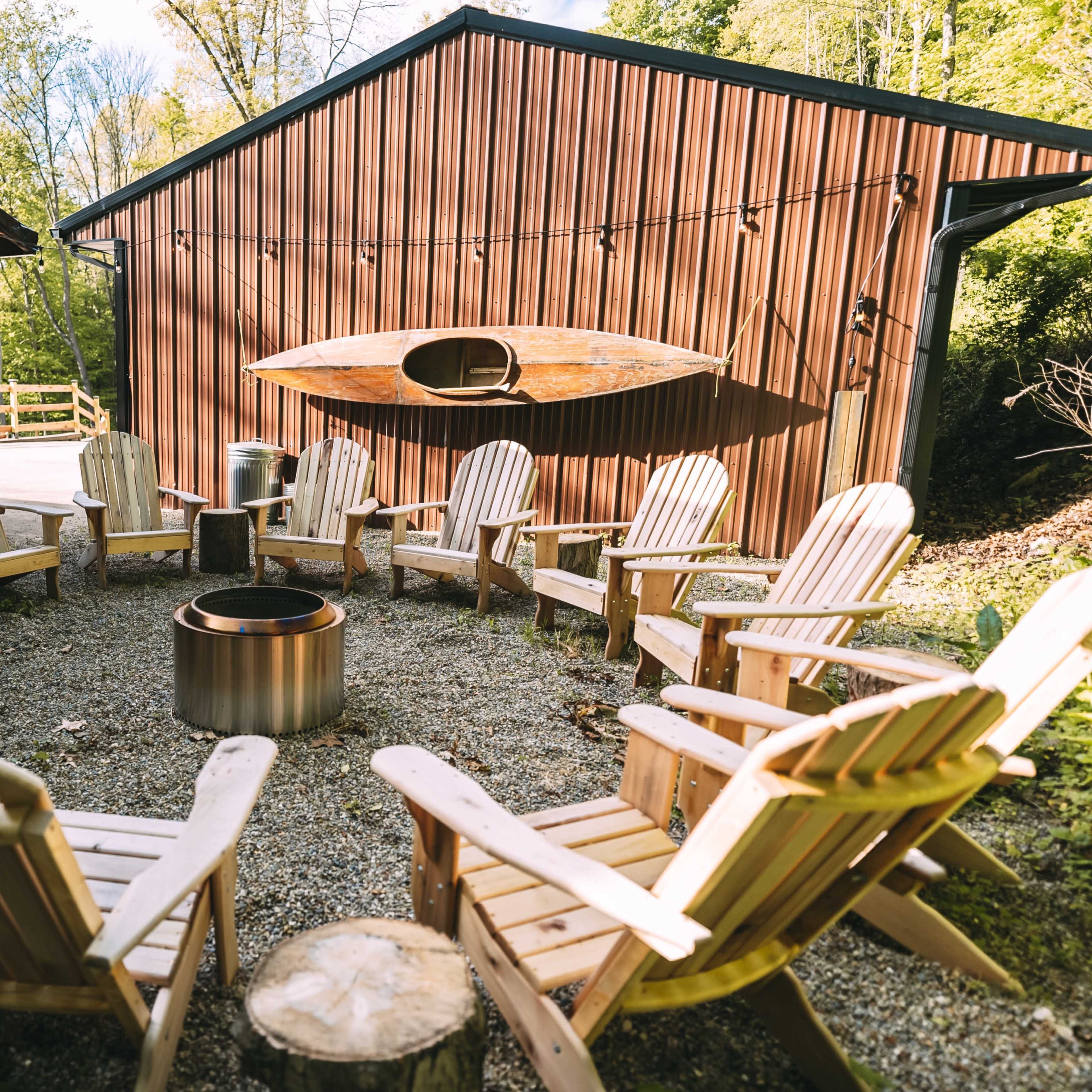 A circular arrangement of wooden chairs surrounds a fire pit between two brown buildings.
