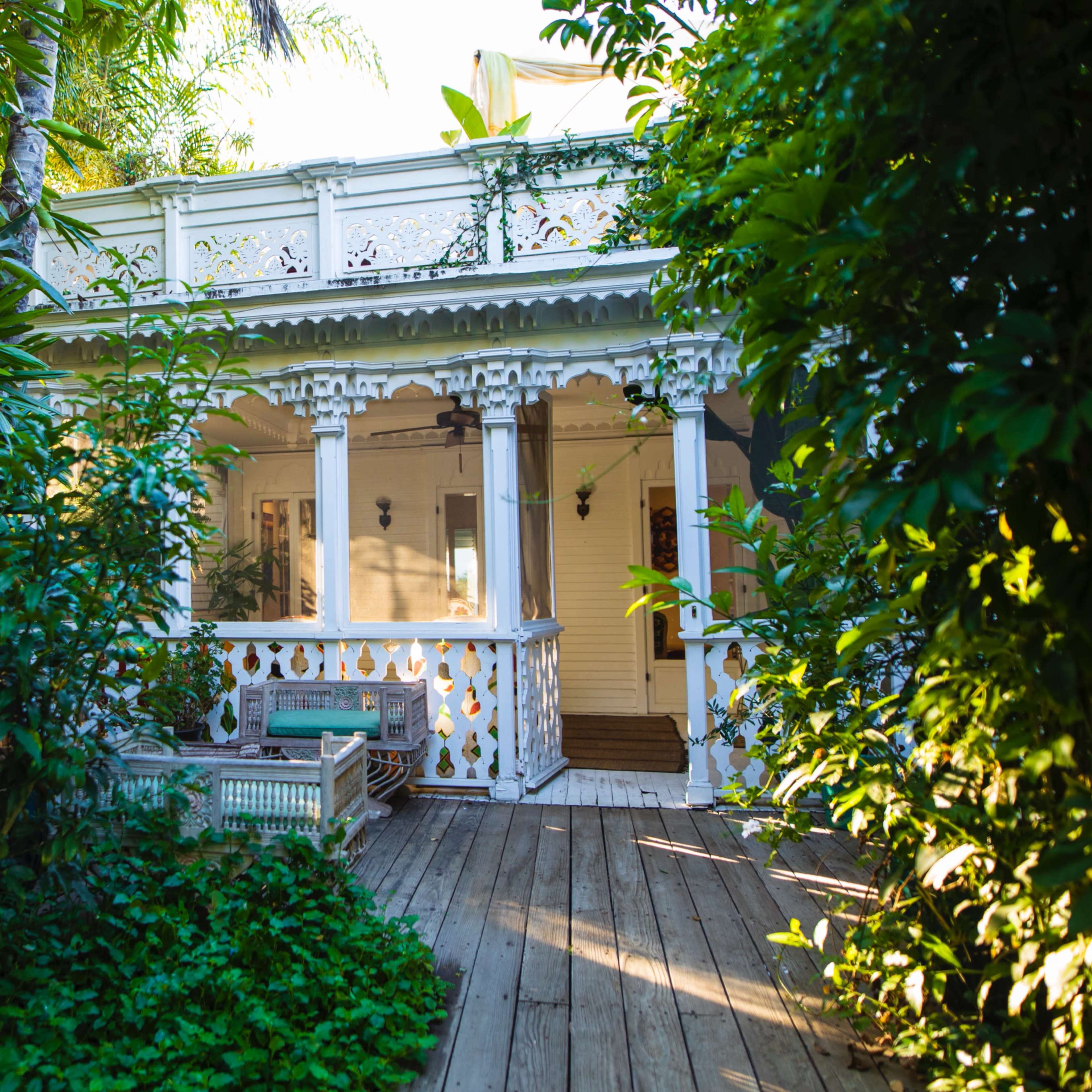 A wooden deck leads to a white, intricately designed house surrounded by lush greenery.