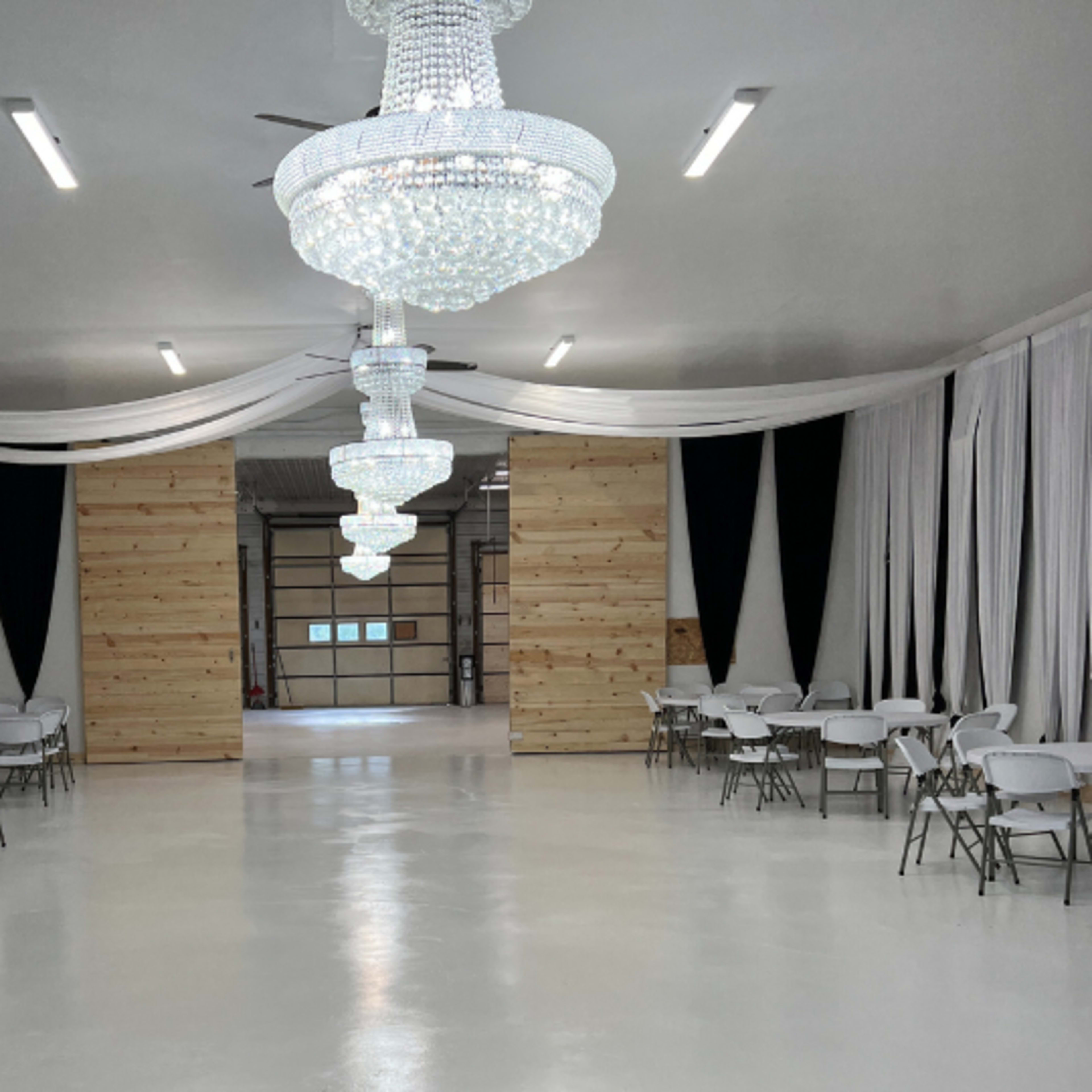 A spacious, well-lit interior with white walls, elegant chandeliers, and rows of folding chairs set up around white tables, framed by fabric drapes.
