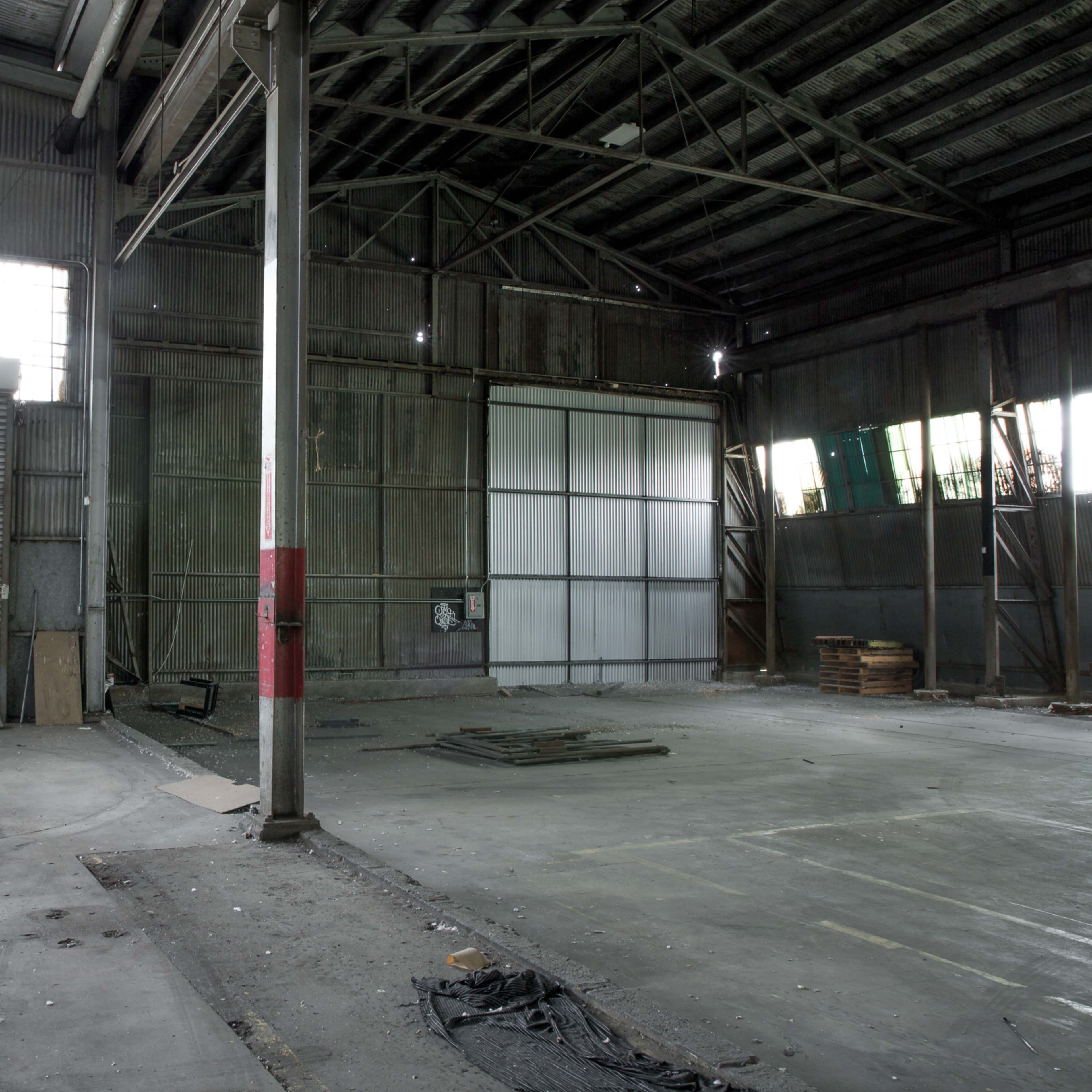 An empty industrial warehouse with large open spaces, a concrete floor, and metal walls featuring a closed roll-up door and a large white panel.