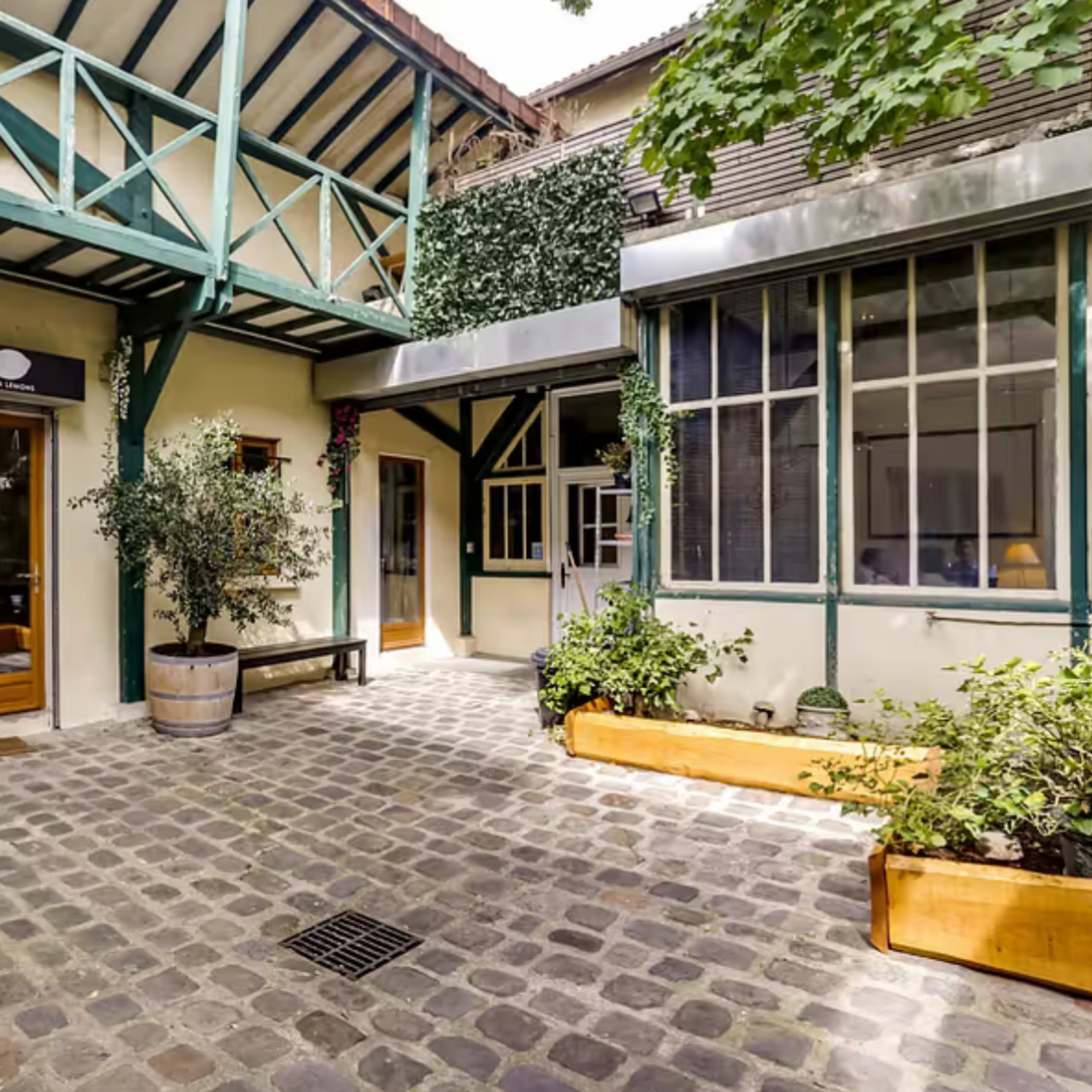 A courtyard featuring cobblestone flooring, a wooden bench, and buildings with large windows, surrounded by greenery.