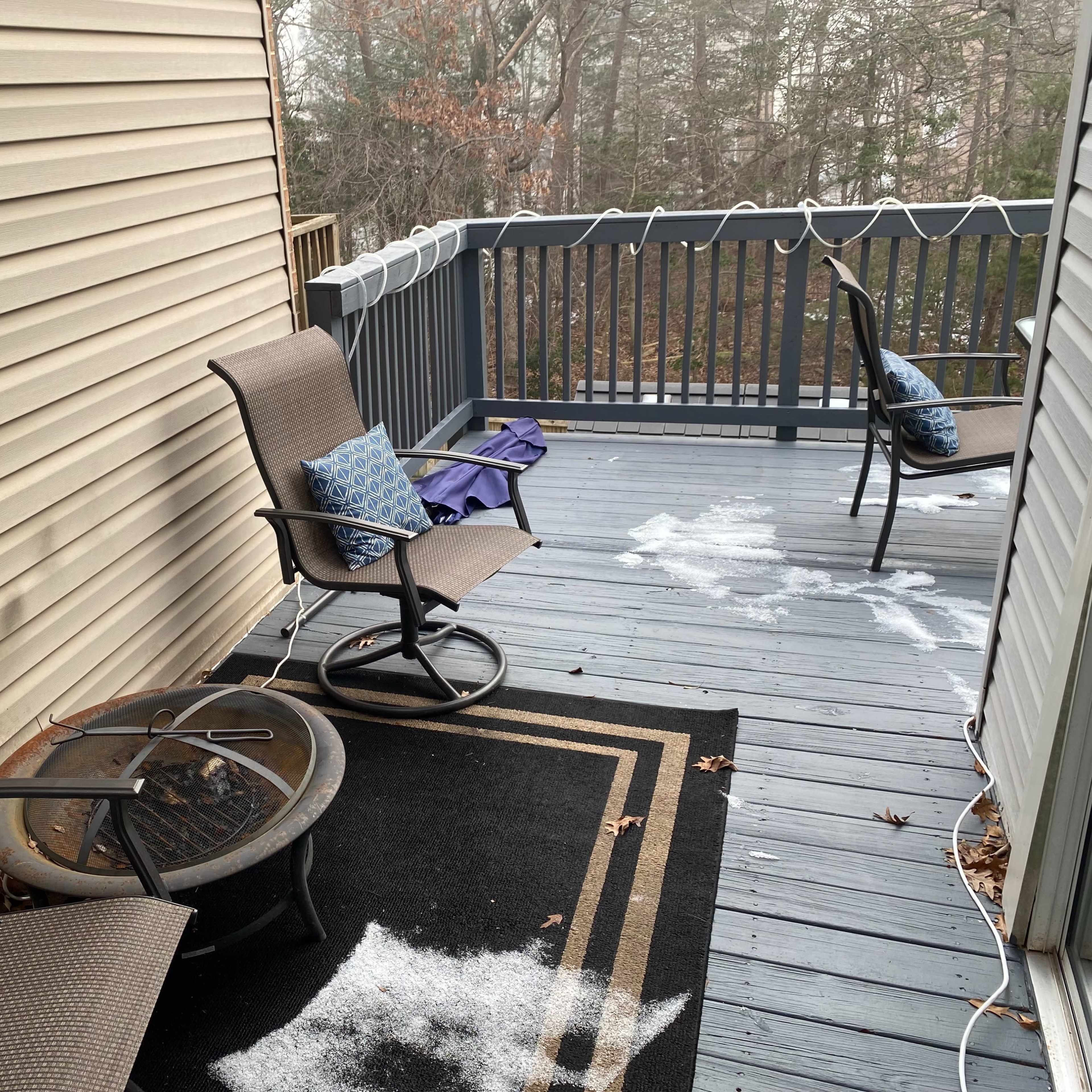 A small deck with two chairs, a table with a patterned rug, and patches of snow scattered on the floor.