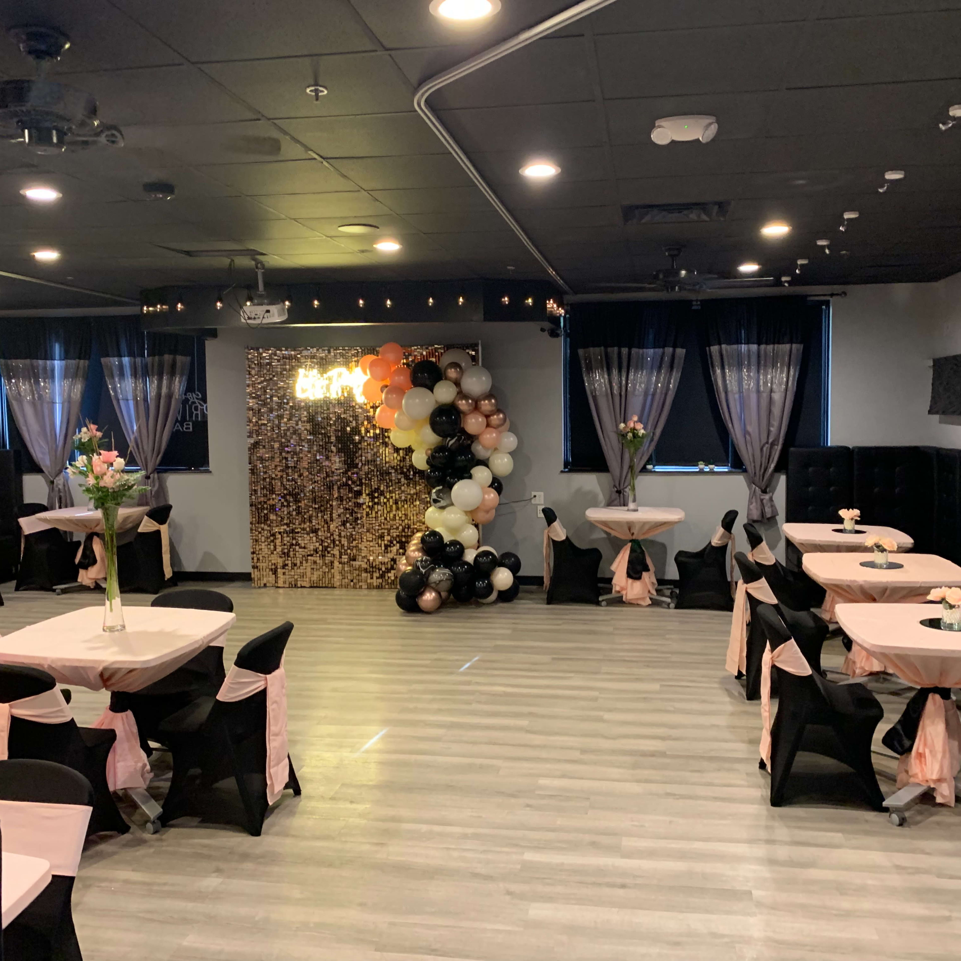 A decorated event space with tables covered in black and pink cloths, surrounded by black chairs, and features a balloon backdrop.