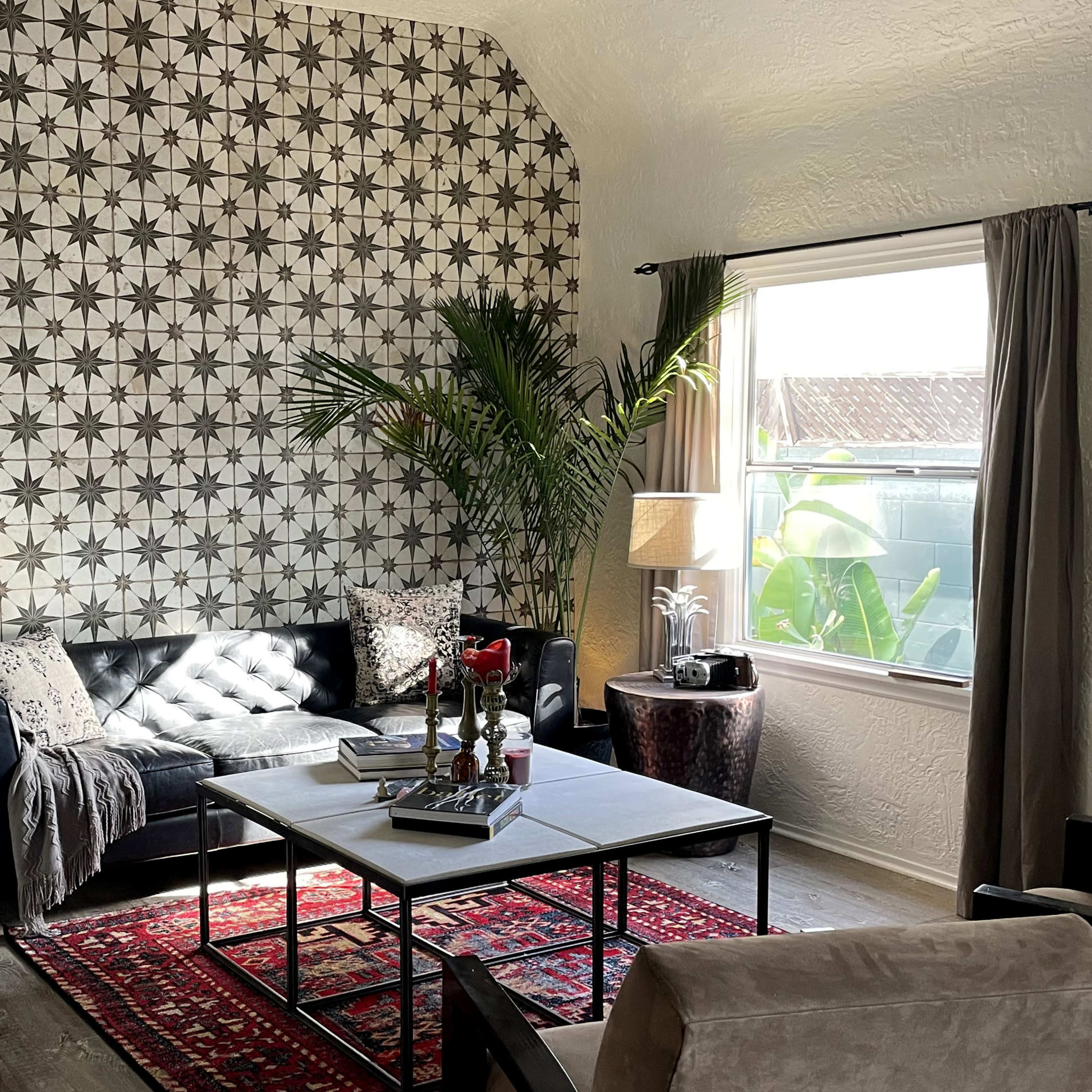 A living room with a patterned wall, a black sofa adorned with a blanket, a coffee table with decor, and a potted plant beside a window.