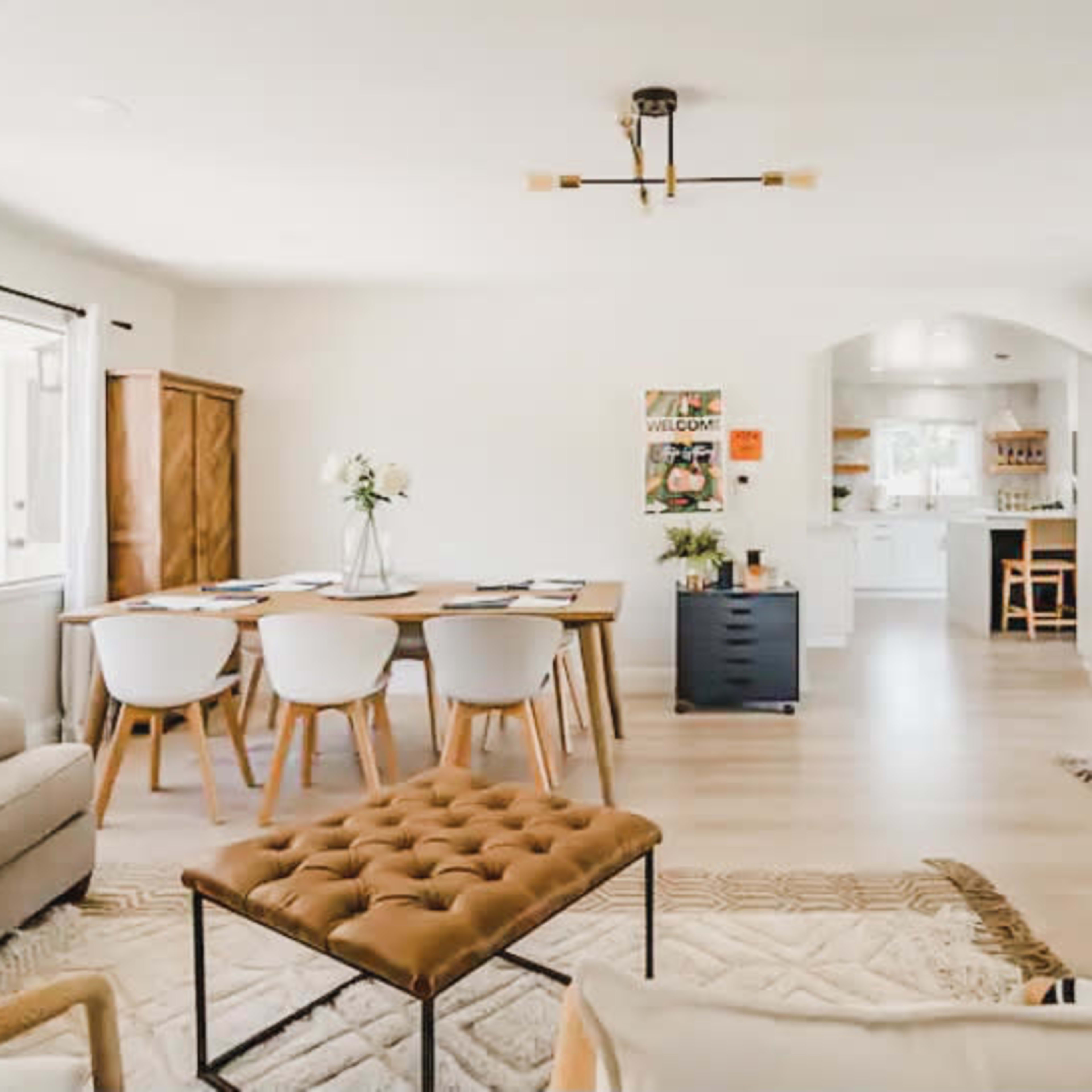 A bright, open living space with a dining area featuring a wooden table and white chairs, leading to a kitchen in the background.