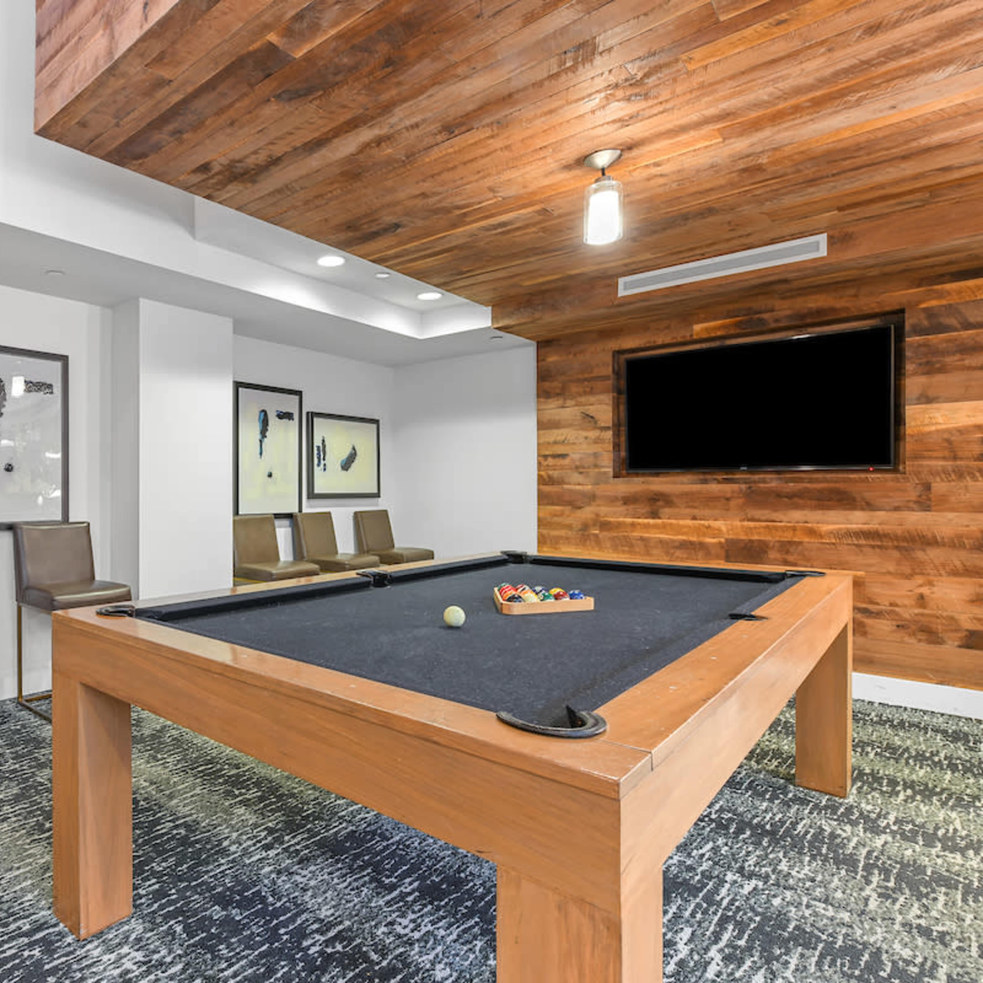A game room featuring a pool table, bar stools, and framed artwork on the walls, with wooden paneling and a television mounted in the corner.