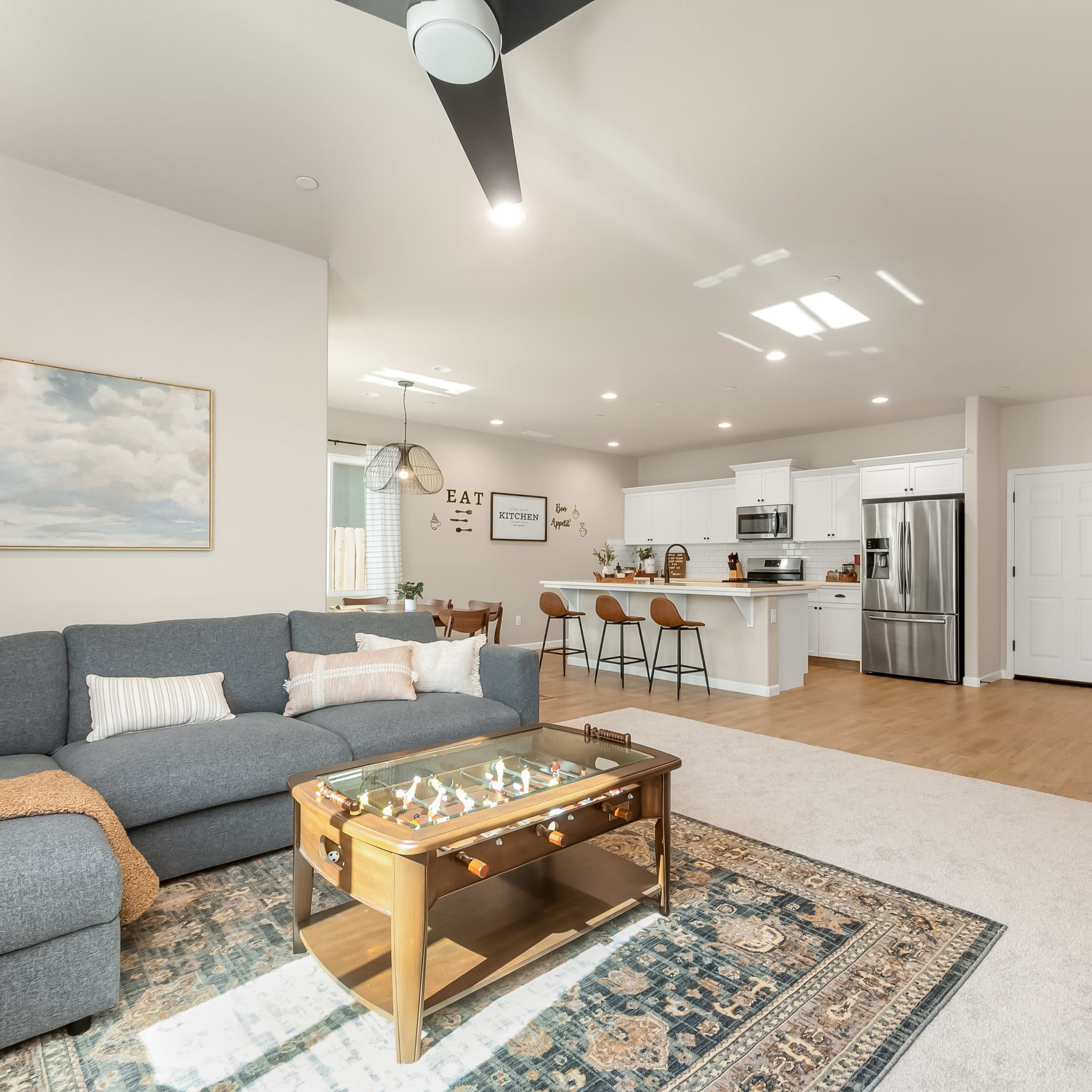 A modern living room features a gray sectional sofa, a glass-top coffee table, and an open kitchen area with bar seating.