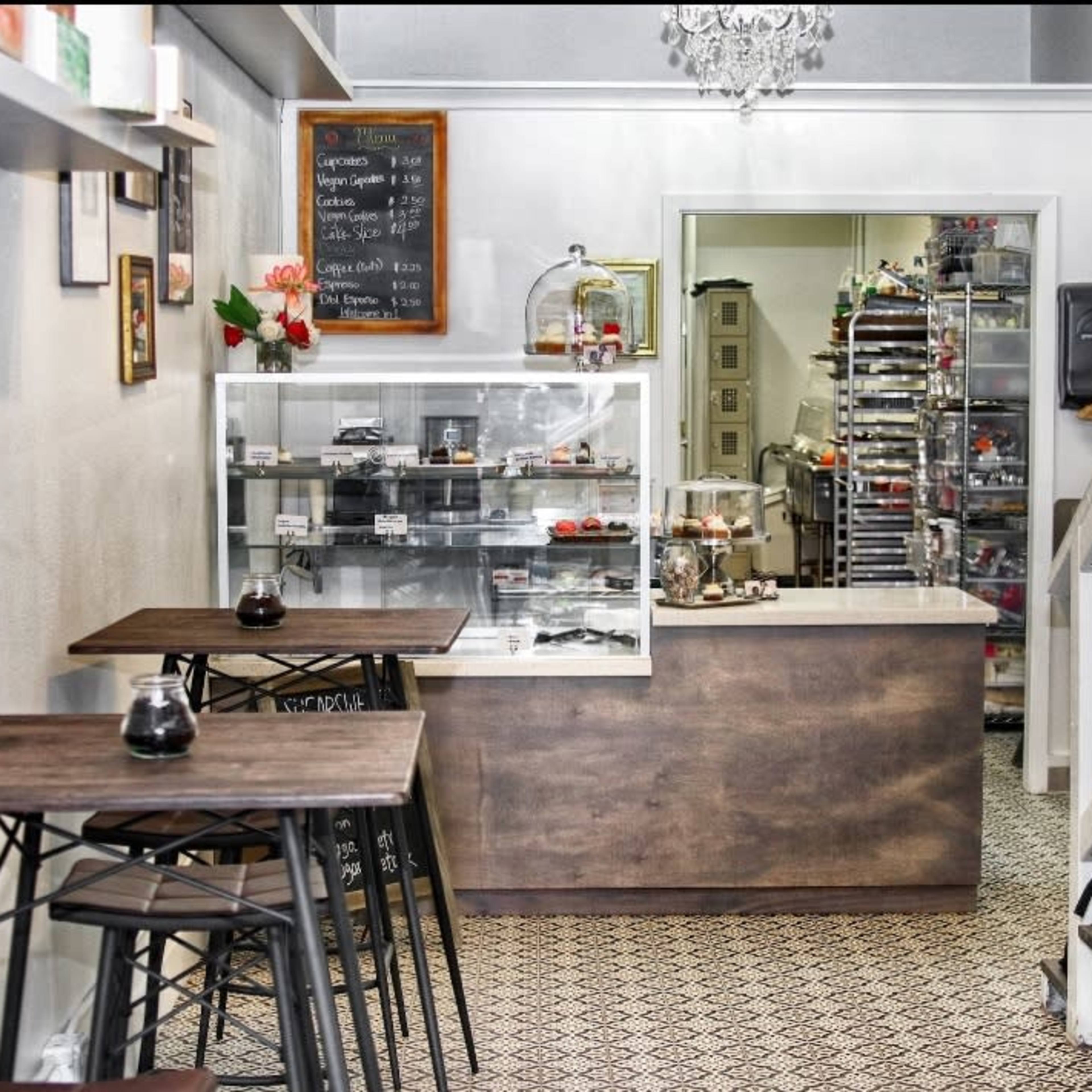 A small café interior with two wooden tables, a display counter filled with pastries, and a staircase leading to an upper level.