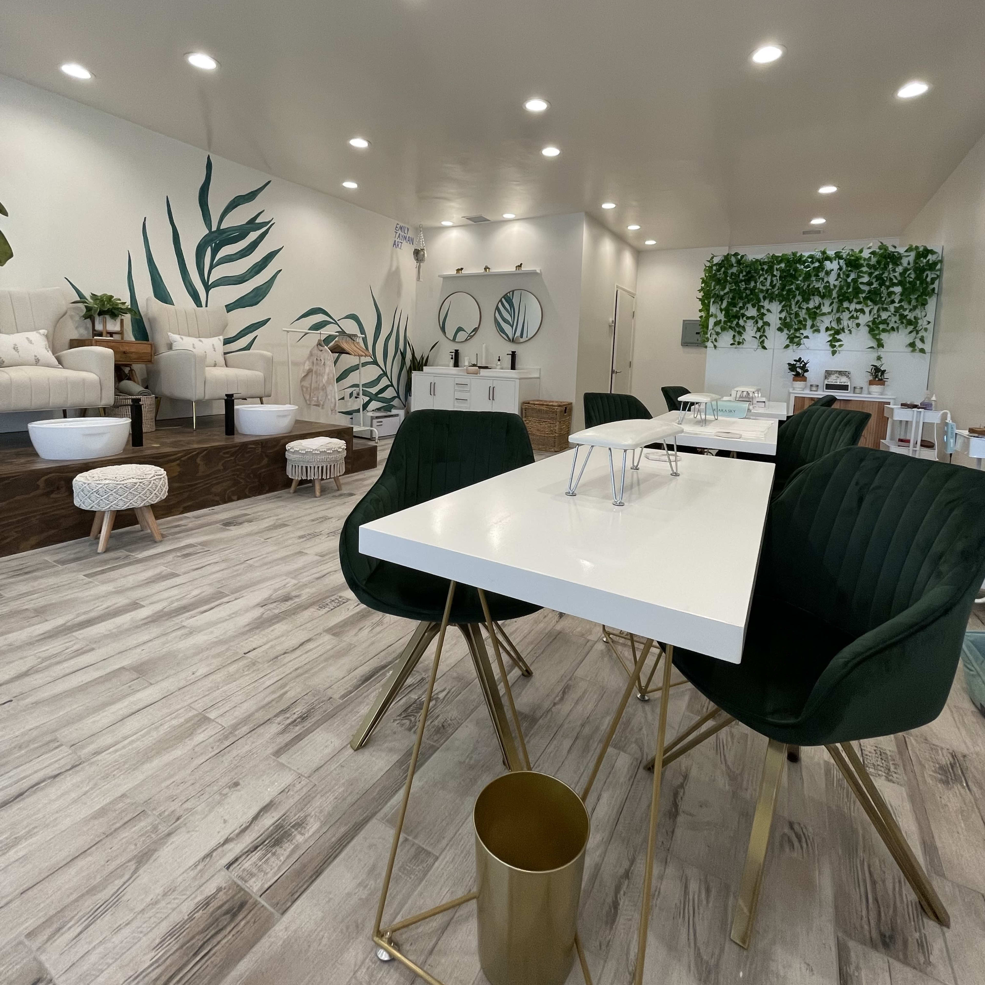 A modern salon interior with green accent chairs, white tables, and a decorated wall featuring plants.