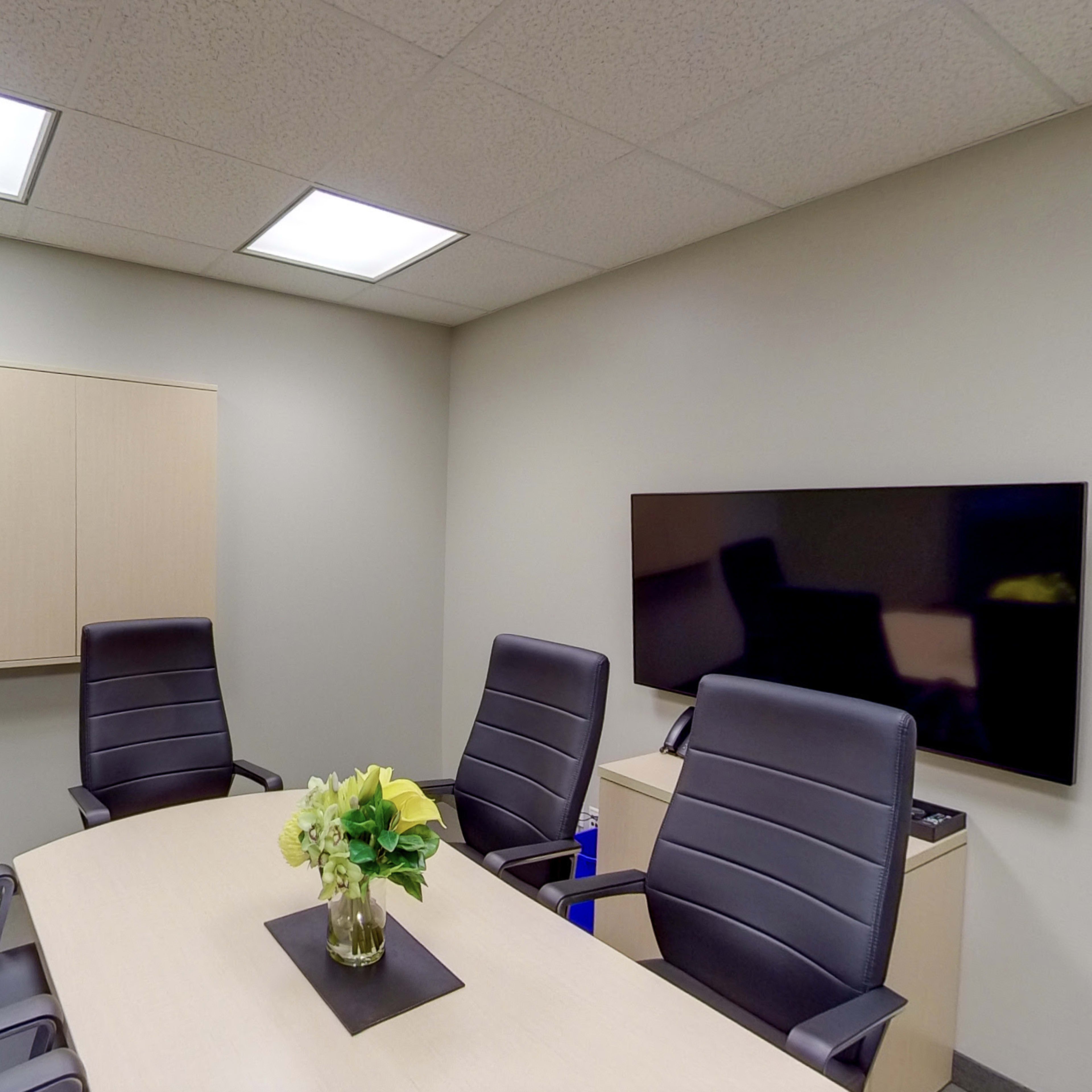 A conference room features a rectangular table surrounded by four black leather chairs, with a small vase of flowers in the center and a wall-mounted TV on one side.