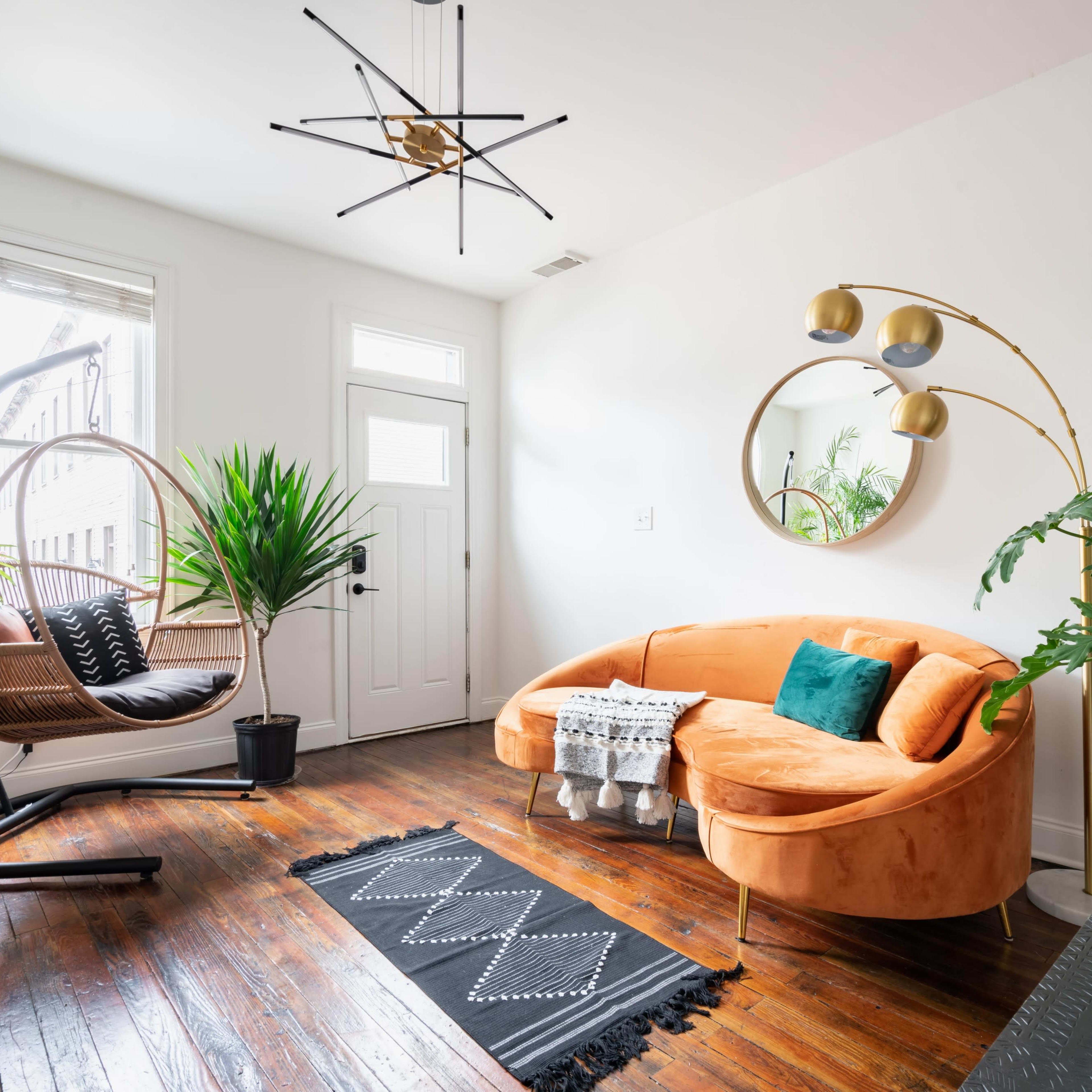 A modern living space featuring an orange curved sofa, a hanging chair, various indoor plants, and a round mirror on a white wall.