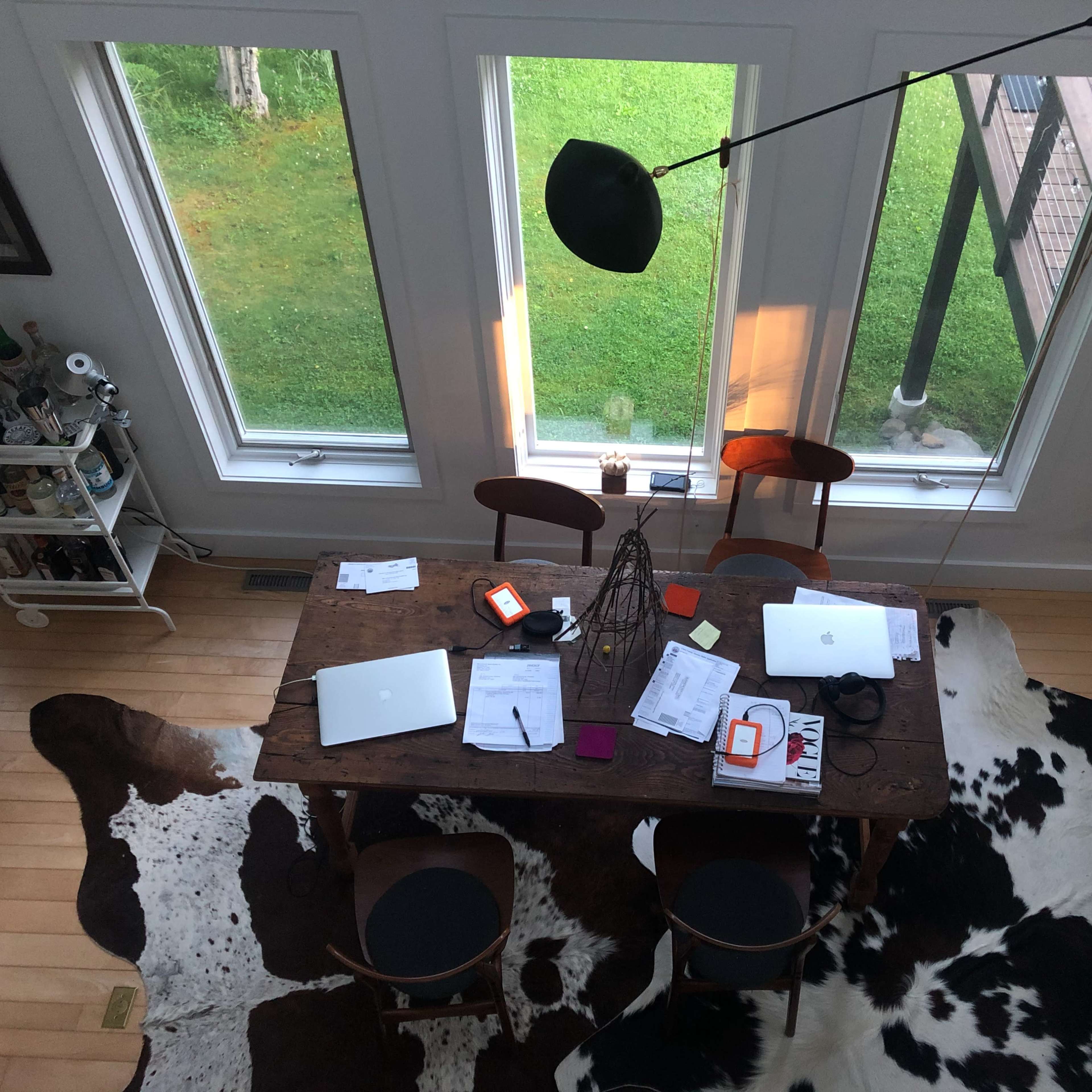 A large wooden table adorned with laptops, papers, and miscellaneous items is surrounded by four chairs on a floor covered with a cowhide rug, beneath three large windows.