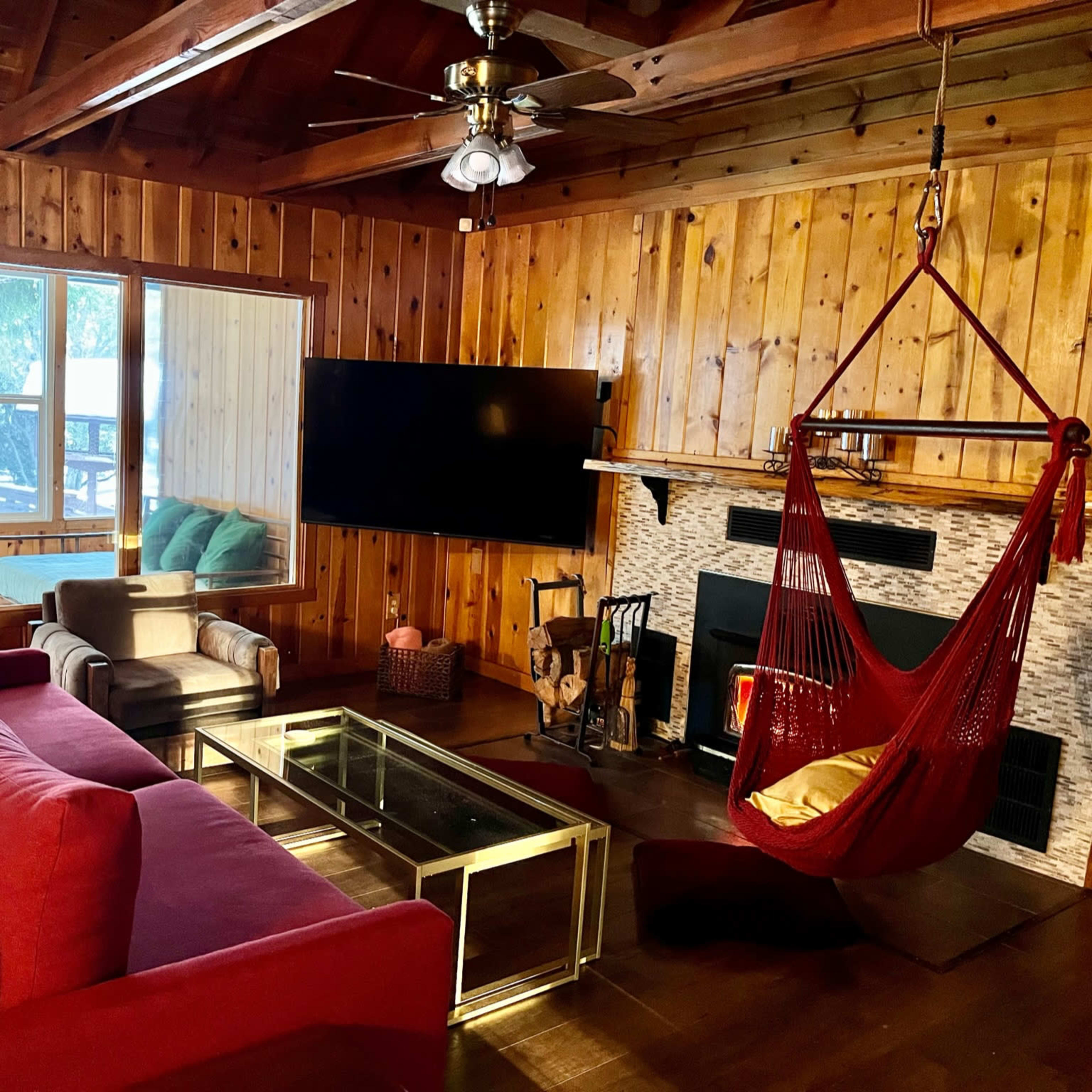 A cozy living room featuring wooden walls, a red sofa, a glass coffee table, and a hanging chair beside a fireplace.