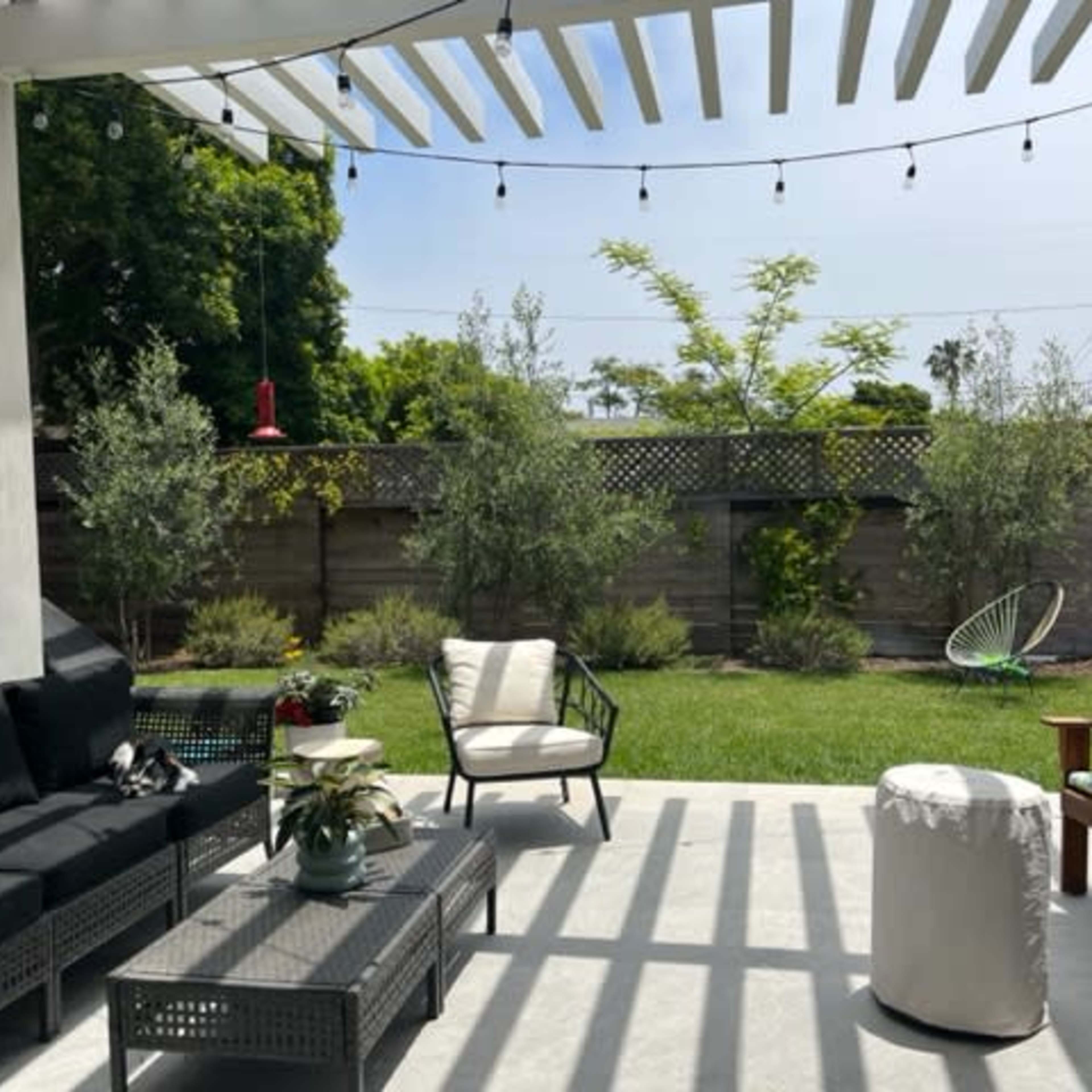 A patio area features a seating arrangement with black furniture, a small table, and a white ottoman, surrounded by greenery and a lawn under a pergola with hanging lights.