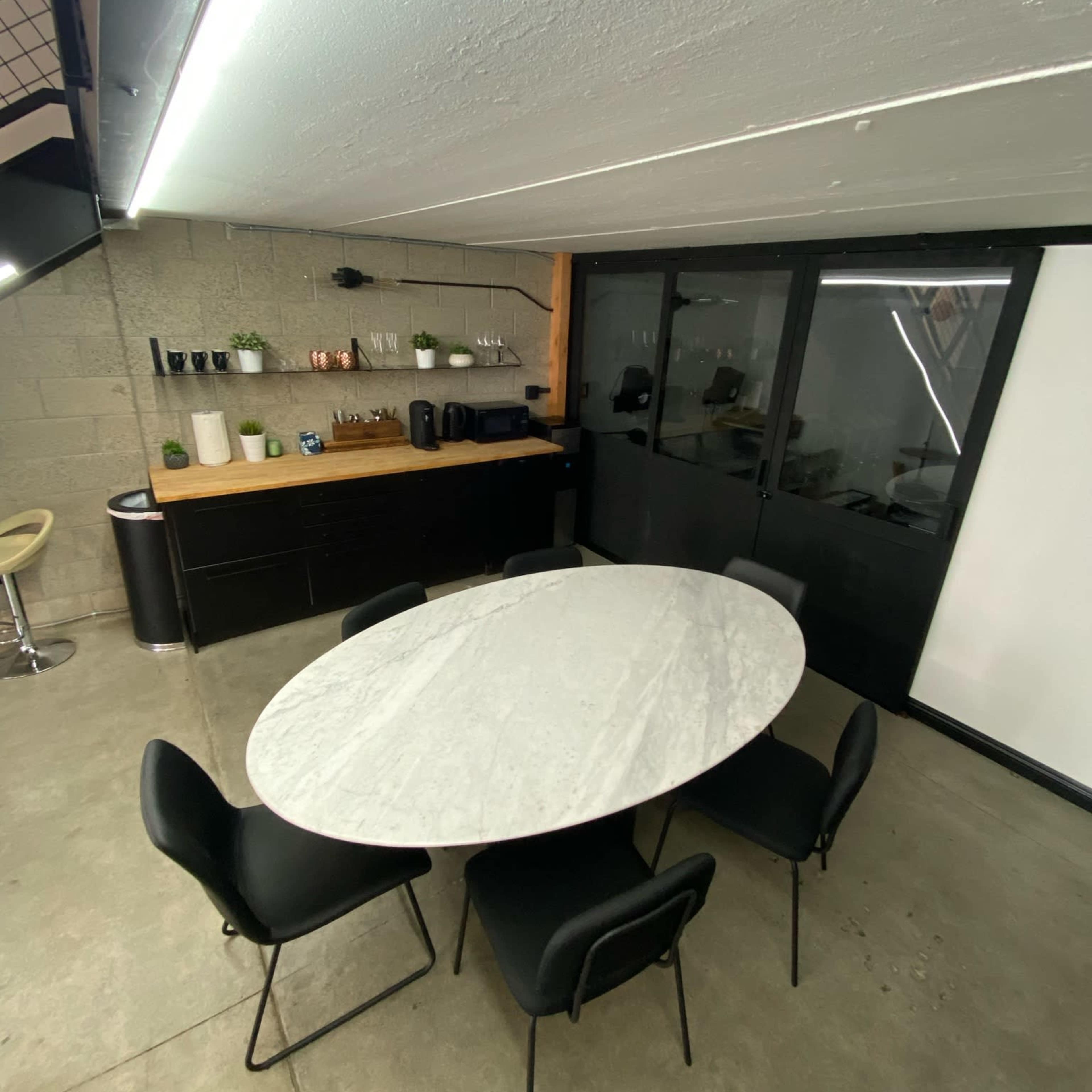 A spacious meeting room features a marble oval table surrounded by six black chairs, with a kitchen area and cabinets in the background.