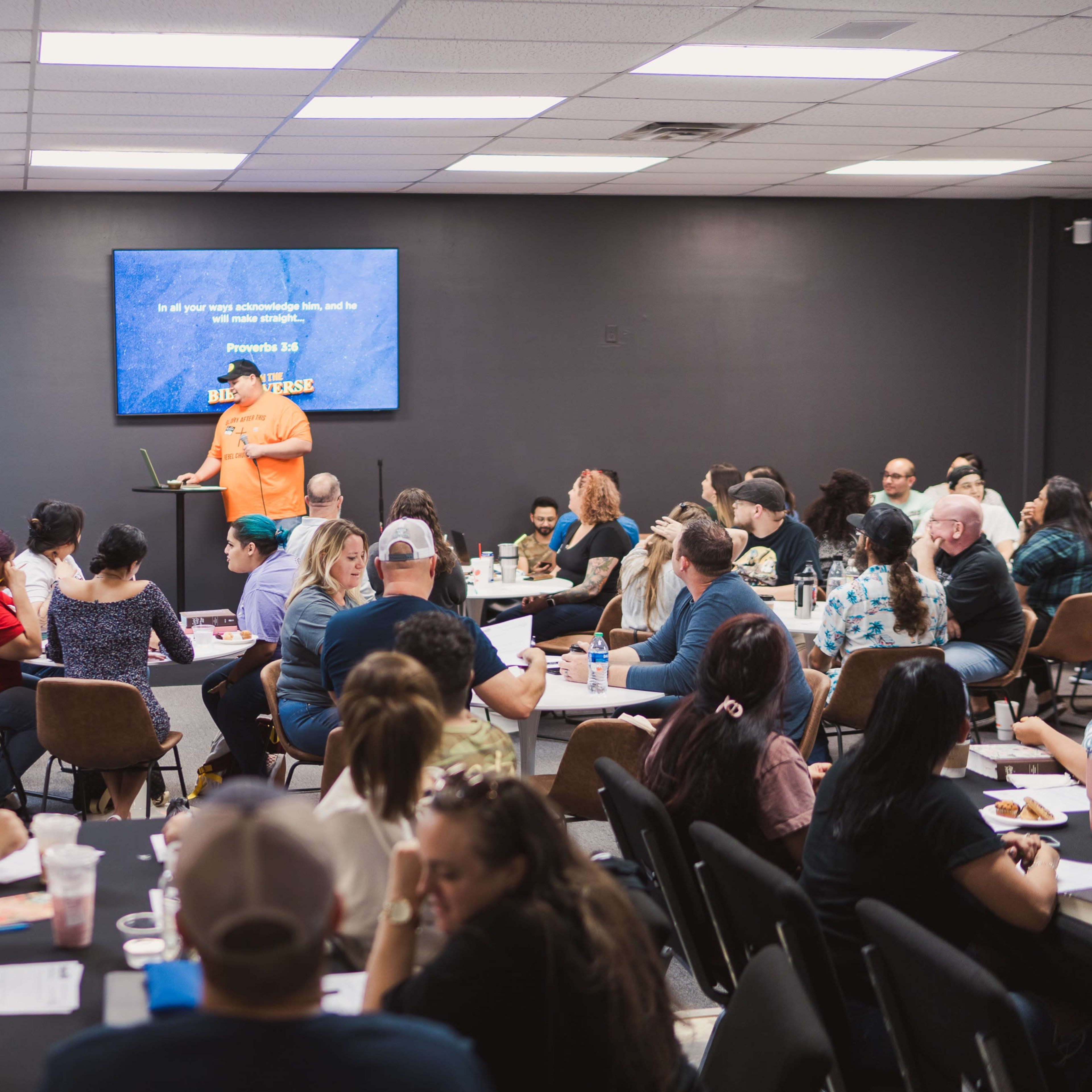 A large group of people sit at tables in a meeting room, attentively listening to a speaker in front of a screen displaying a presentation.
