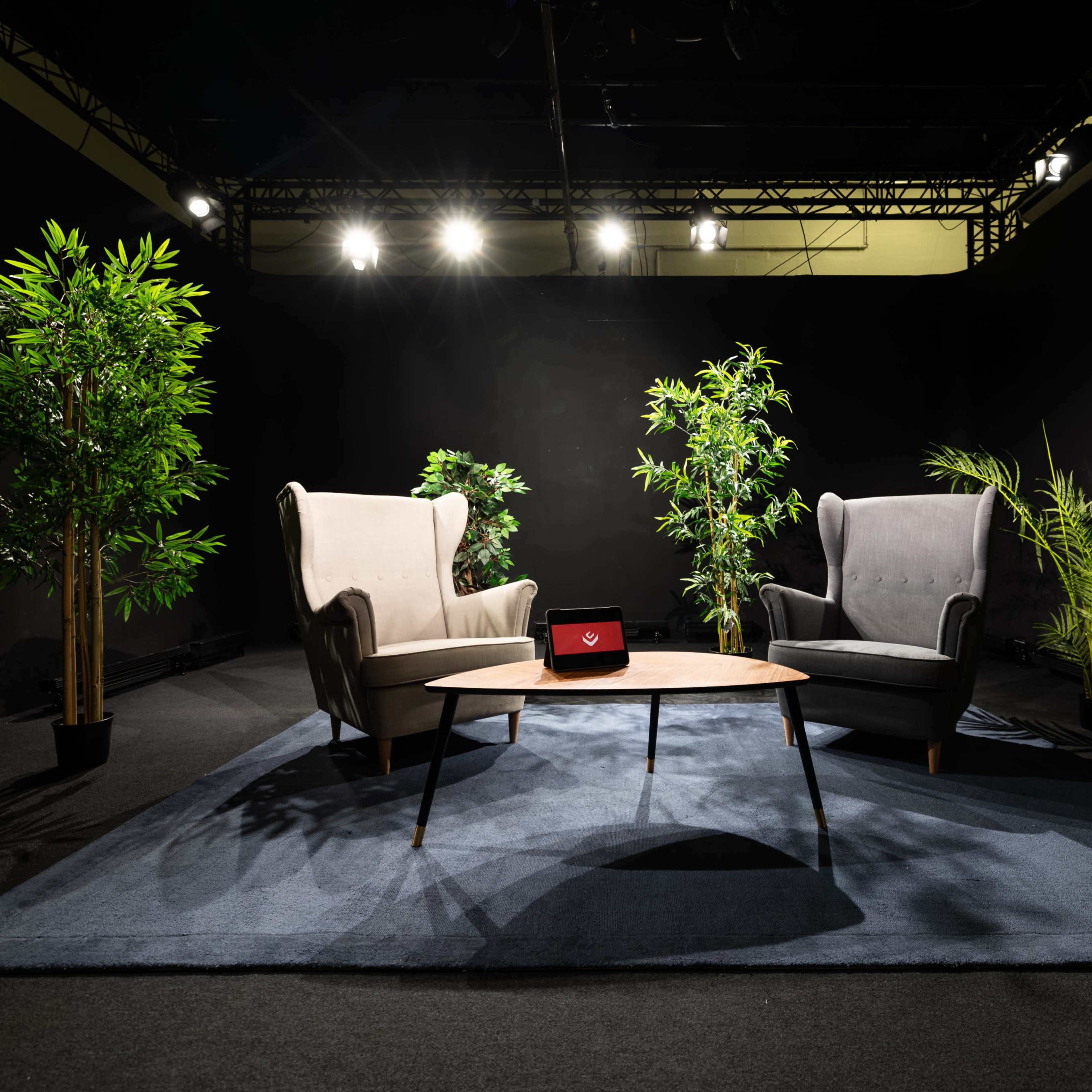 A cozy seating area features two armchairs facing each other, a round table in the center, and potted plants in a dimly lit space.