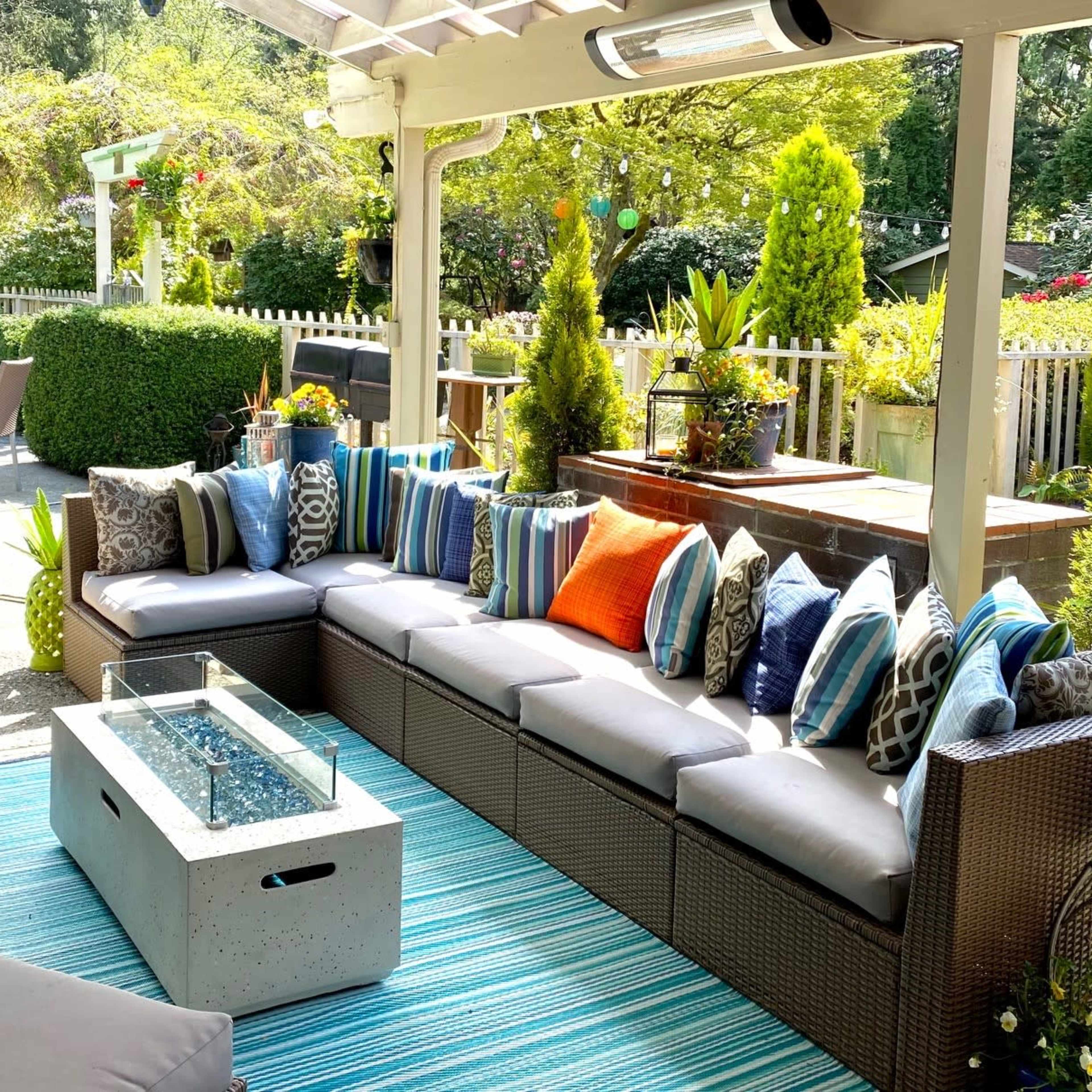 A patio area features a sectional sofa adorned with various throw pillows, a concrete fire table, and a striped outdoor rug, surrounded by greenery and decorative plants.