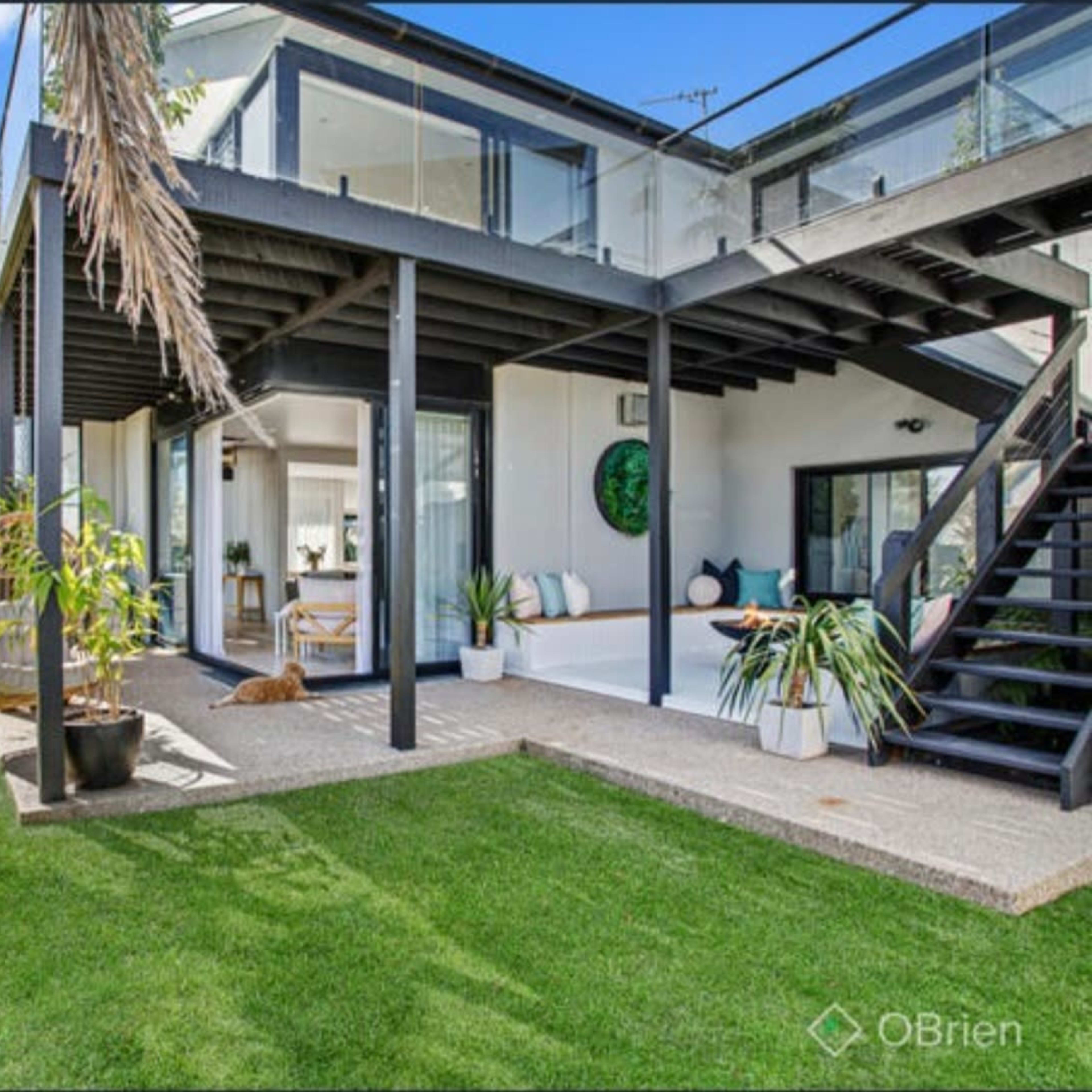 A modern two-story house with large windows, a terrace, and a well-maintained grassy yard surrounded by palm trees.