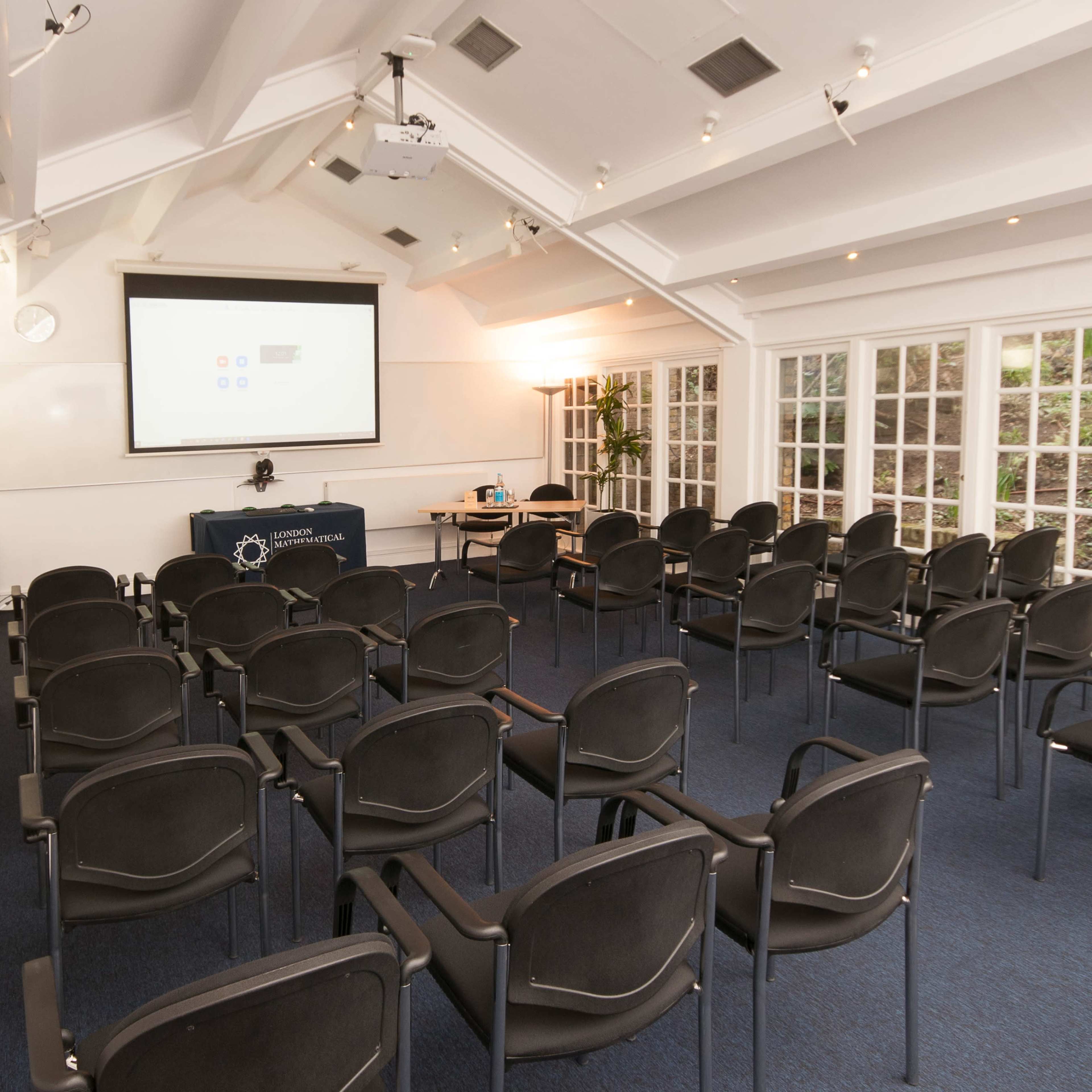 A conference room is arranged with rows of black chairs facing a projection screen and a table at the front.