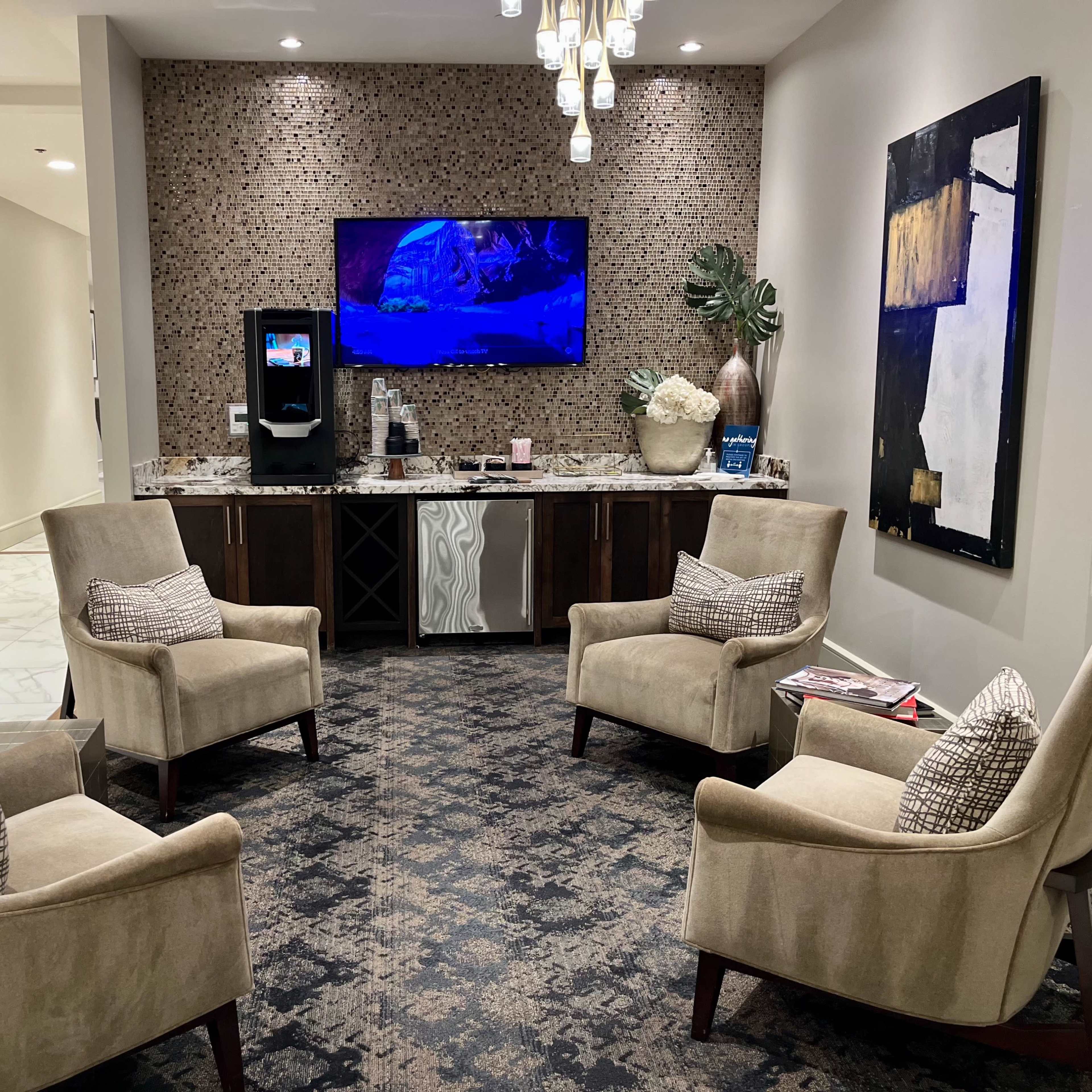 A waiting area with four beige armchairs arranged around a central table, a wall-mounted television, and a refreshment station with decorative elements.