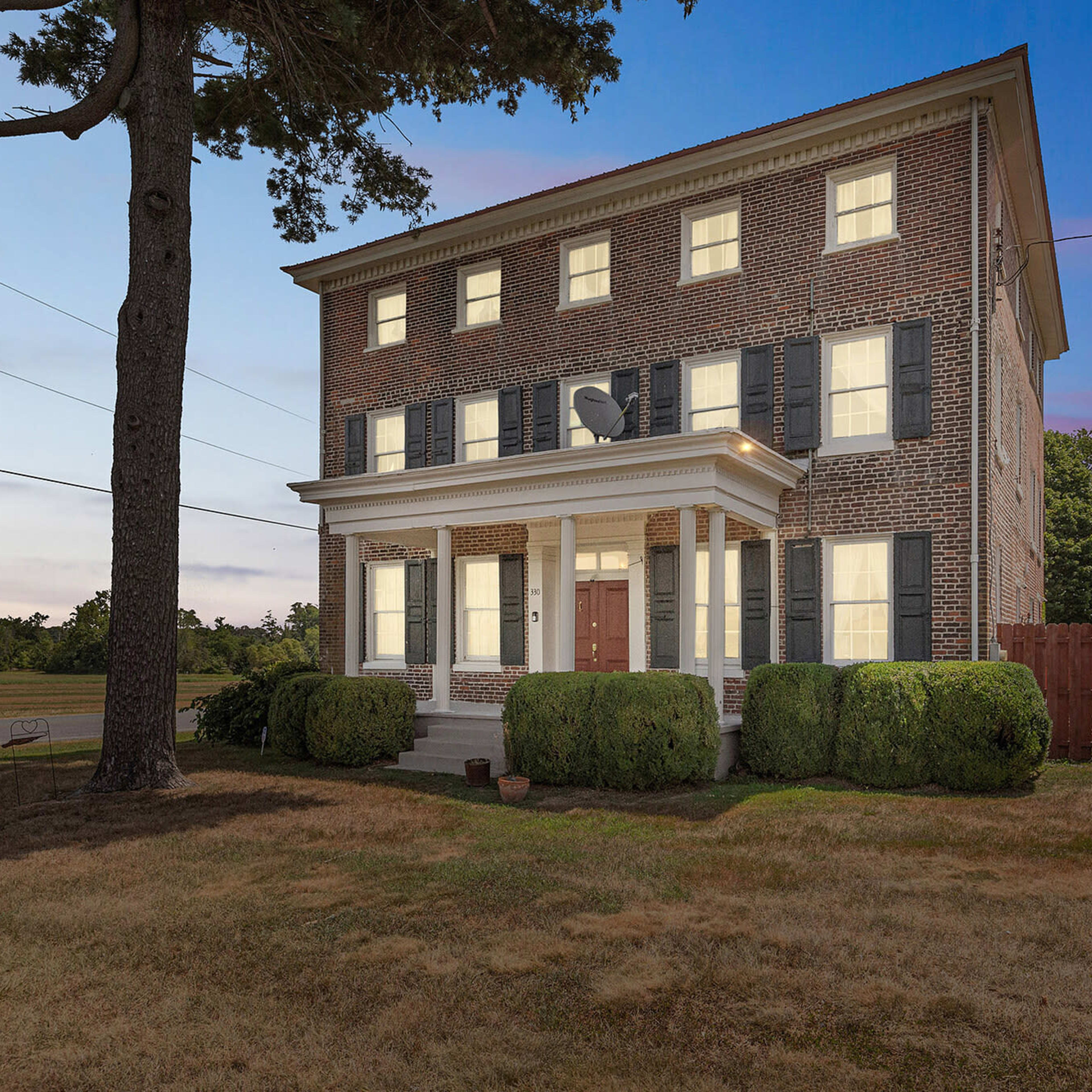 A three-story brick house with white trim and green shrubs is situated near a road under a twilight sky.
