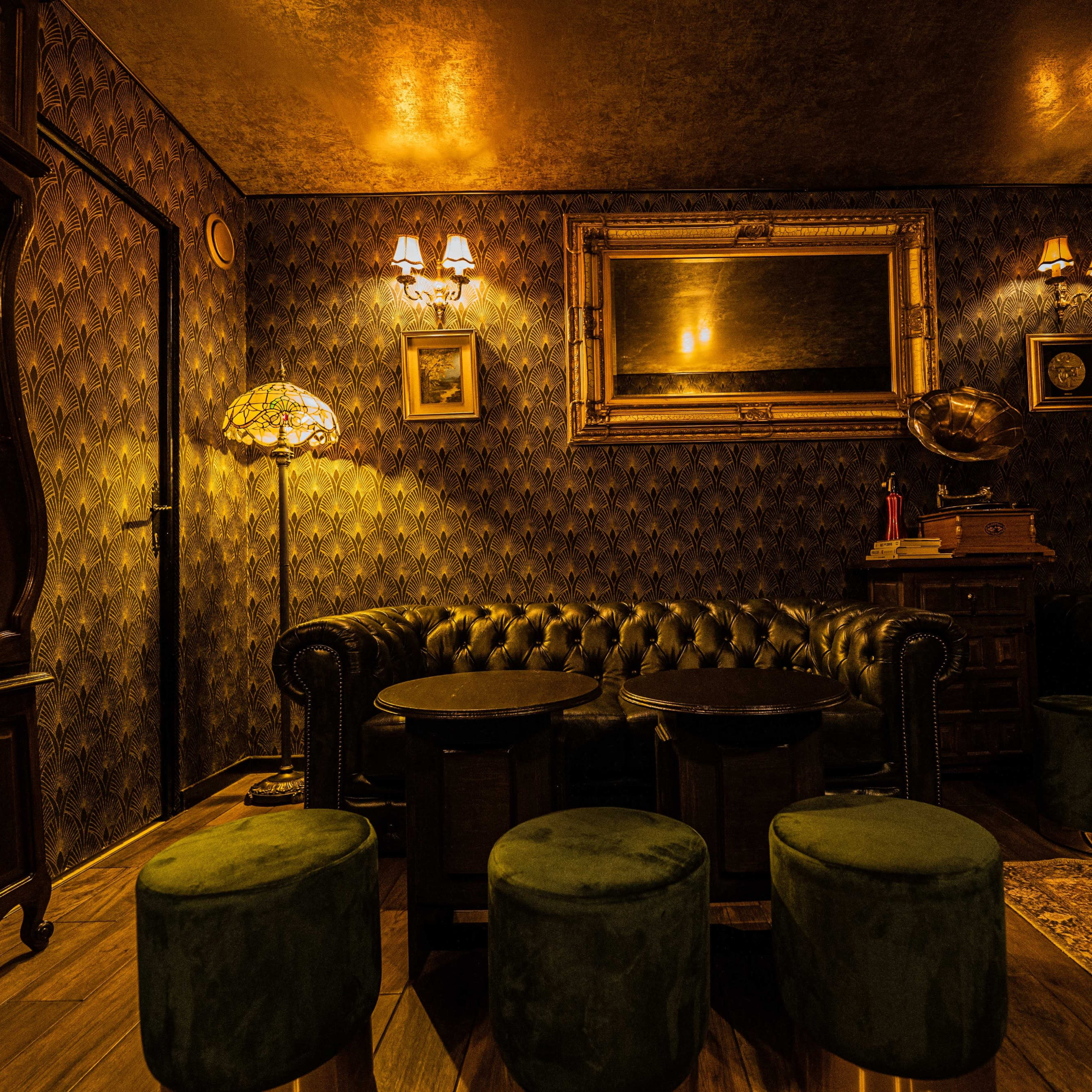 A dimly lit lounge featuring a vintage clock, leather chesterfield sofas, green stools, and decorative wall sconces.