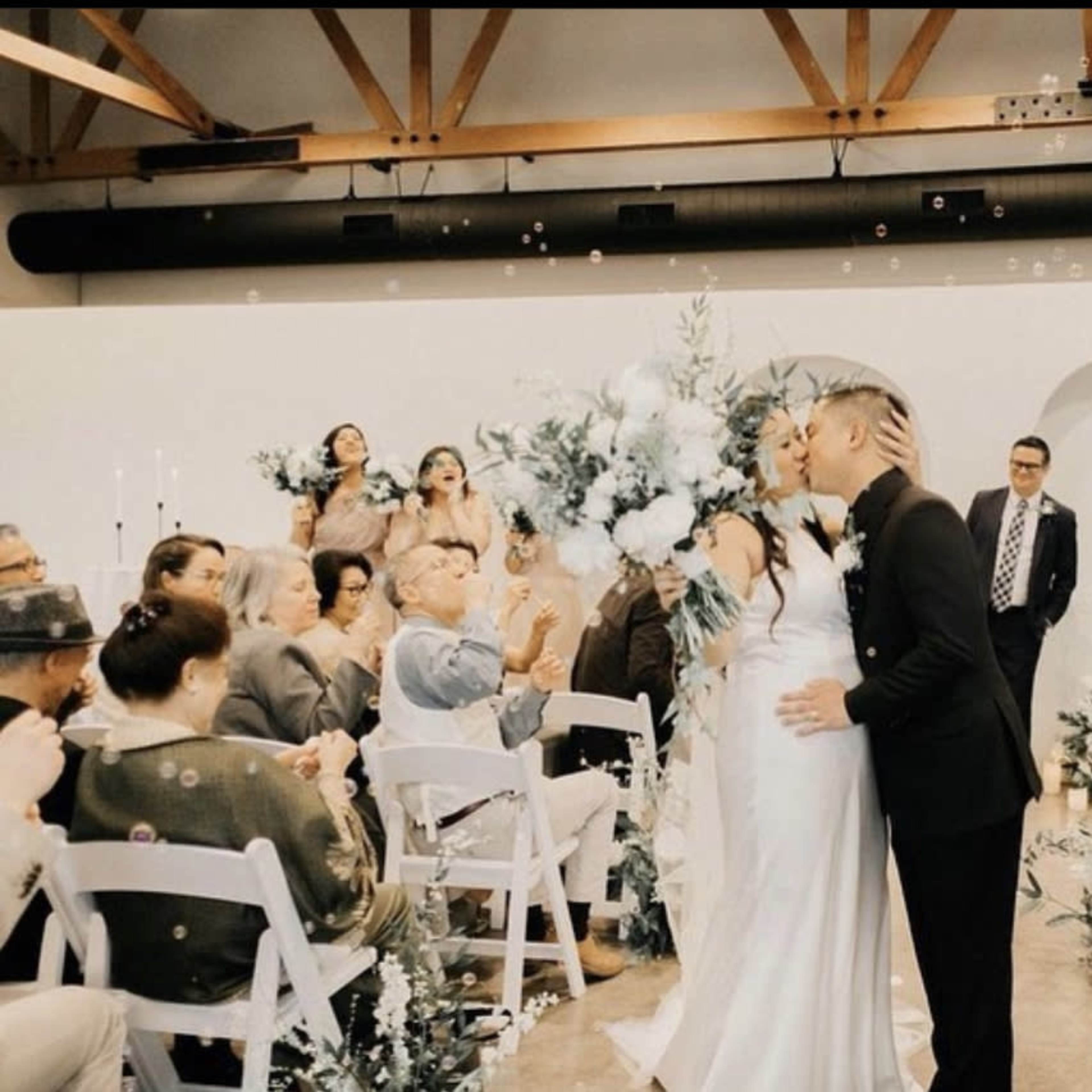 A couple shares a kiss during their wedding ceremony, surrounded by guests and decorated with floral arrangements and bubbles.