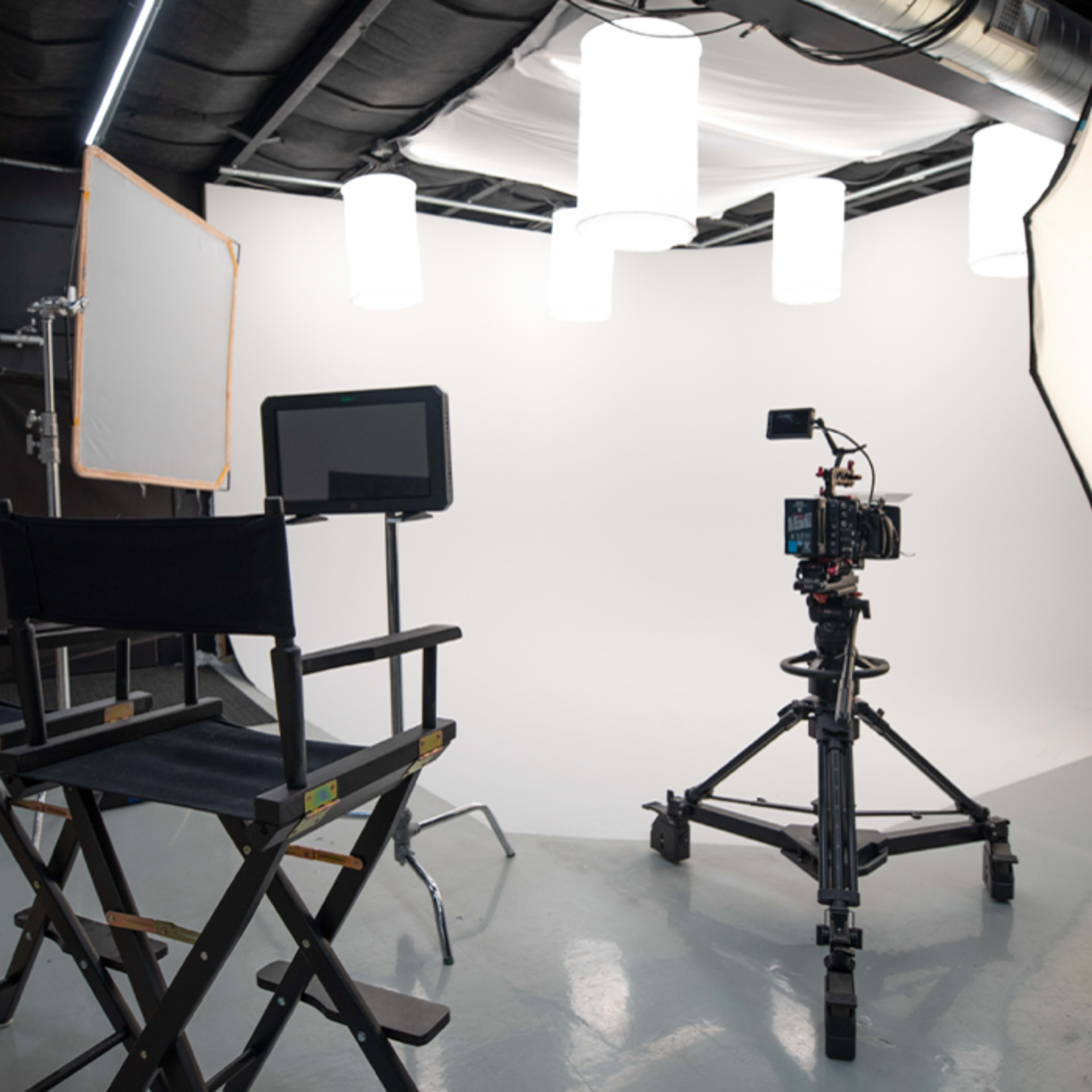 A photography studio equipped with lighting setups, a camera on a tripod, and empty director's chairs facing a white backdrop.