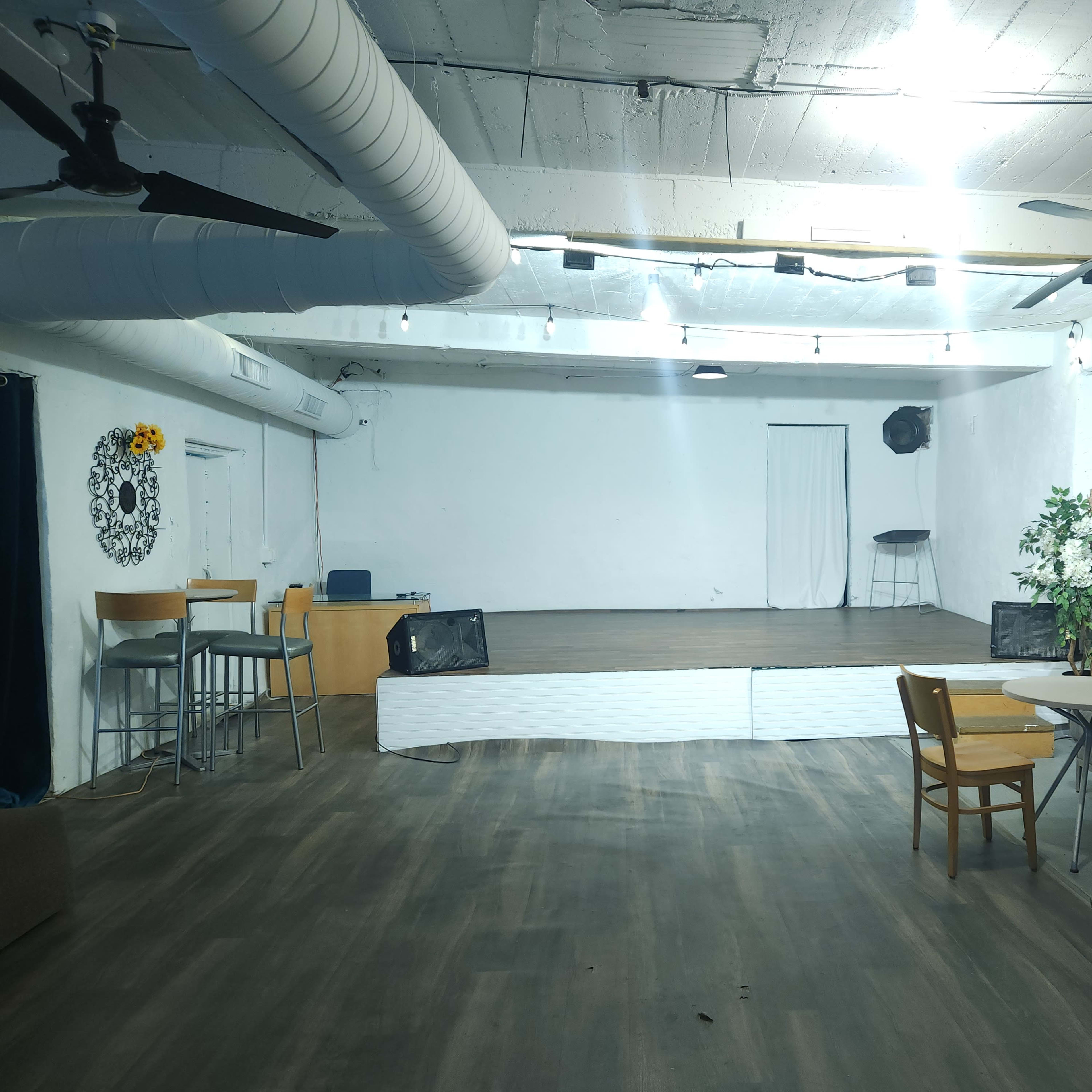 An empty event space with a wooden stage, scattered tables, chairs, and a few decorative elements on the walls.