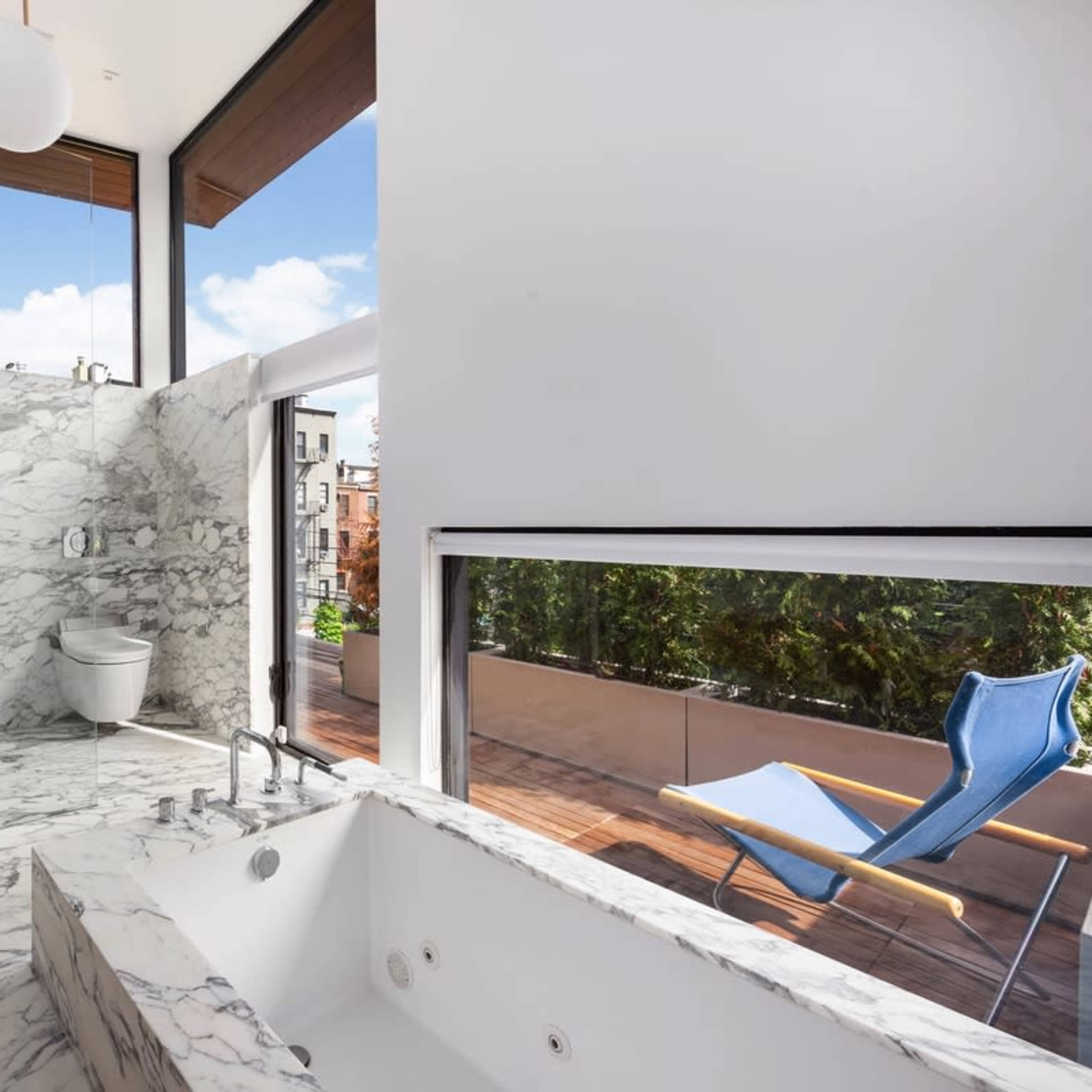 A modern bathroom features marble walls, a freestanding bathtub, and a large window overlooking a wooden balcony with a blue chair.
