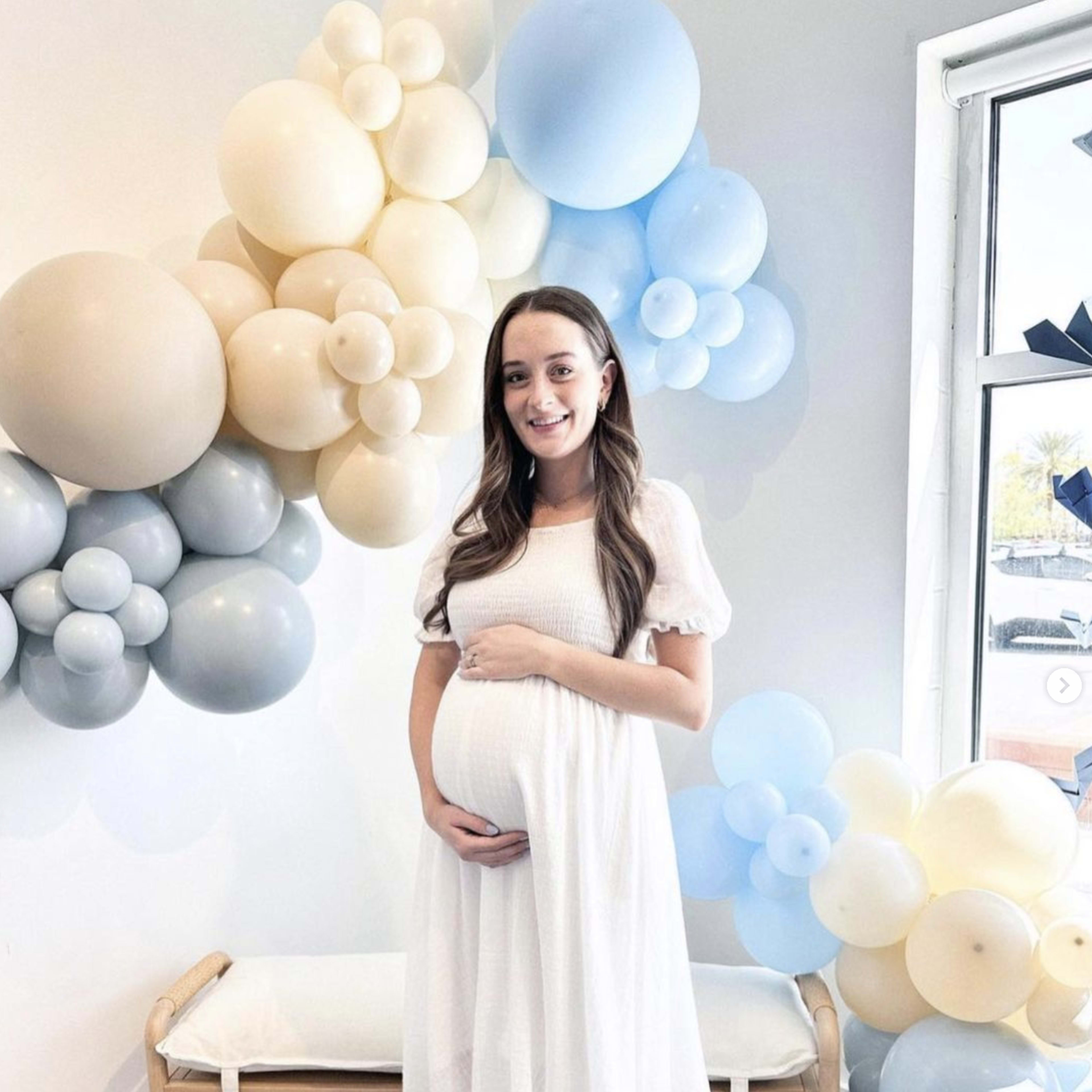 A woman in a white dress stands in front of a wall decorated with clusters of blue and beige balloons, holding her pregnant belly.