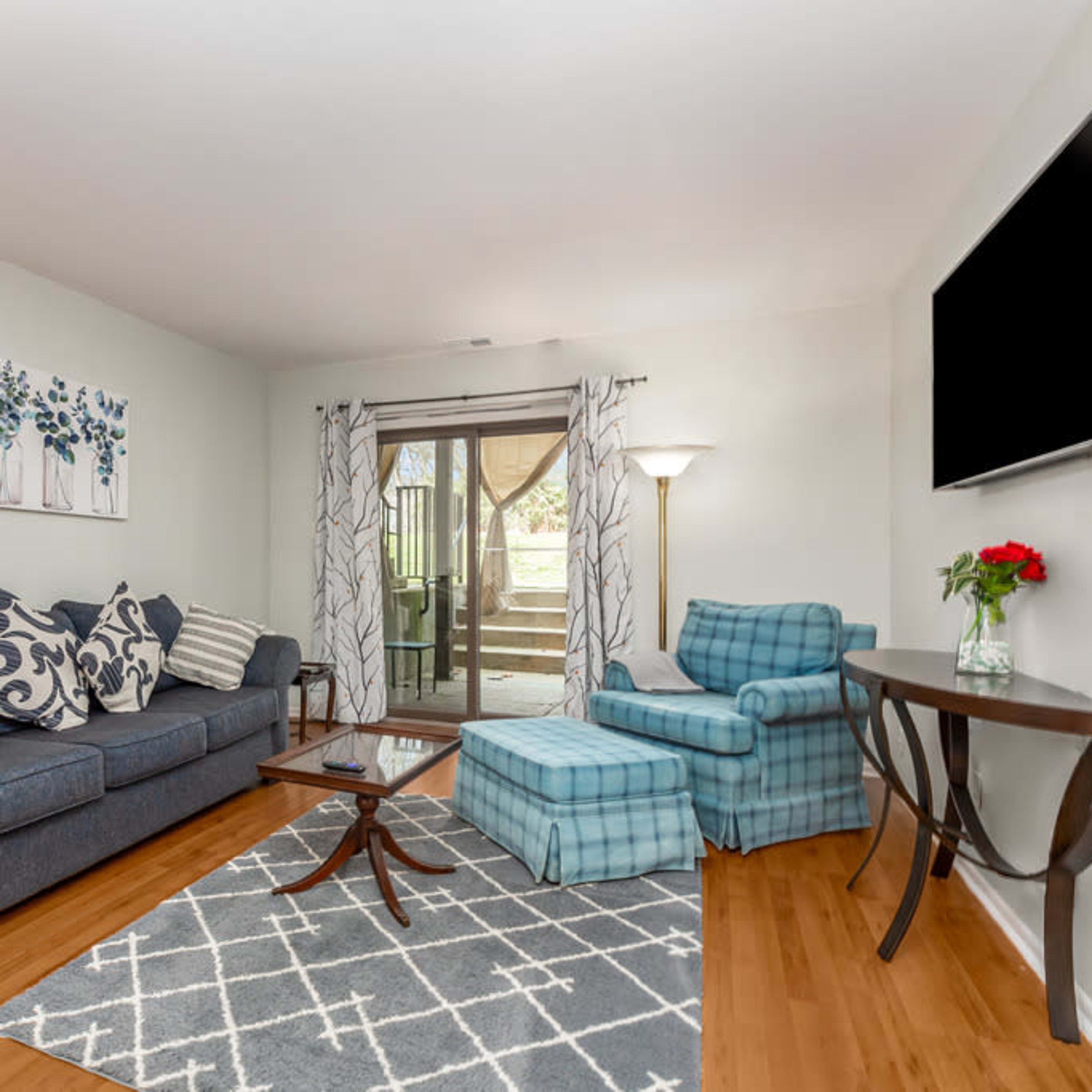 A living room with a blue sofa, a checkered armchair, a coffee table, and a view of an outdoor patio through a sliding door.