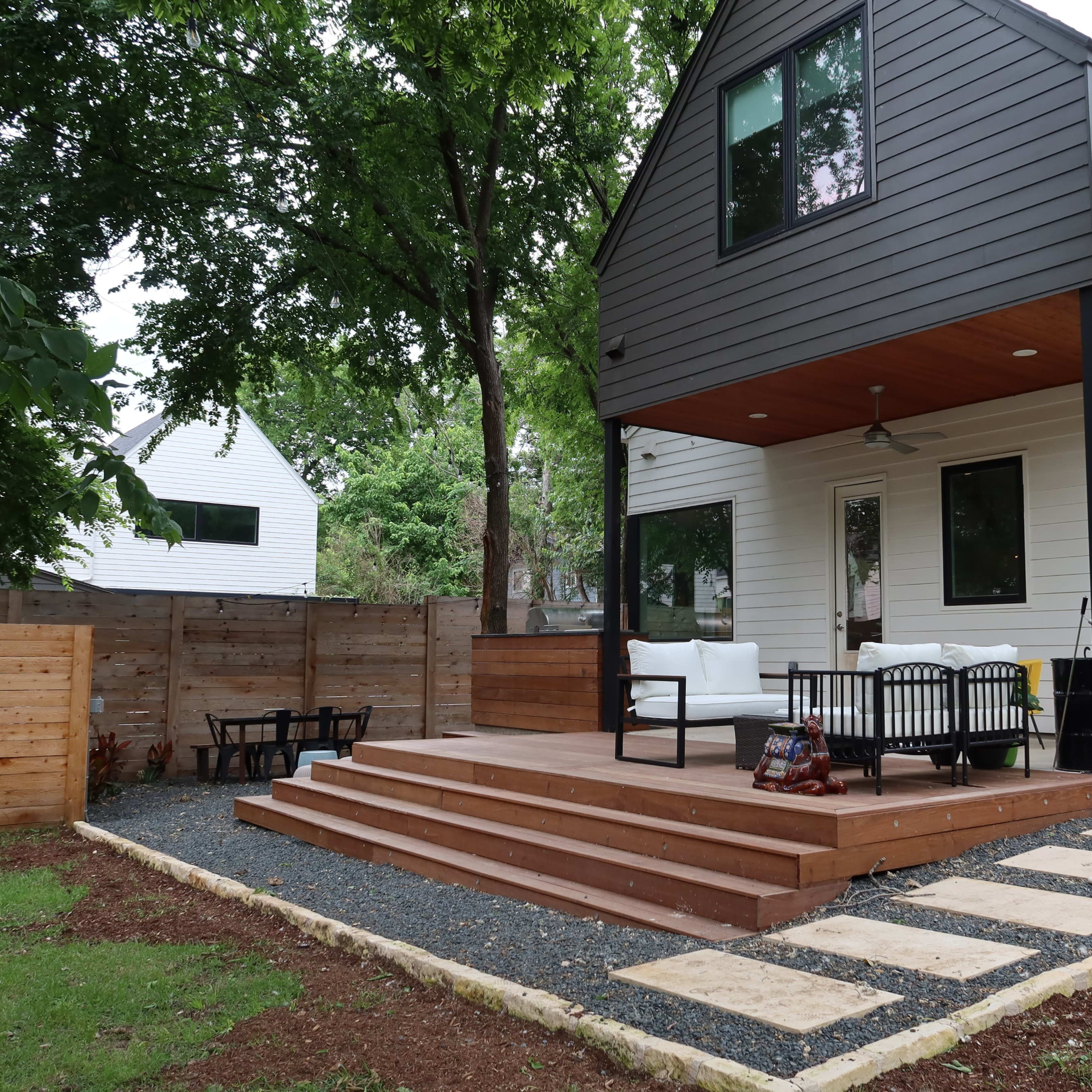 A modern house with a wooden deck and outdoor seating area, surrounded by a fenced yard and landscaping.