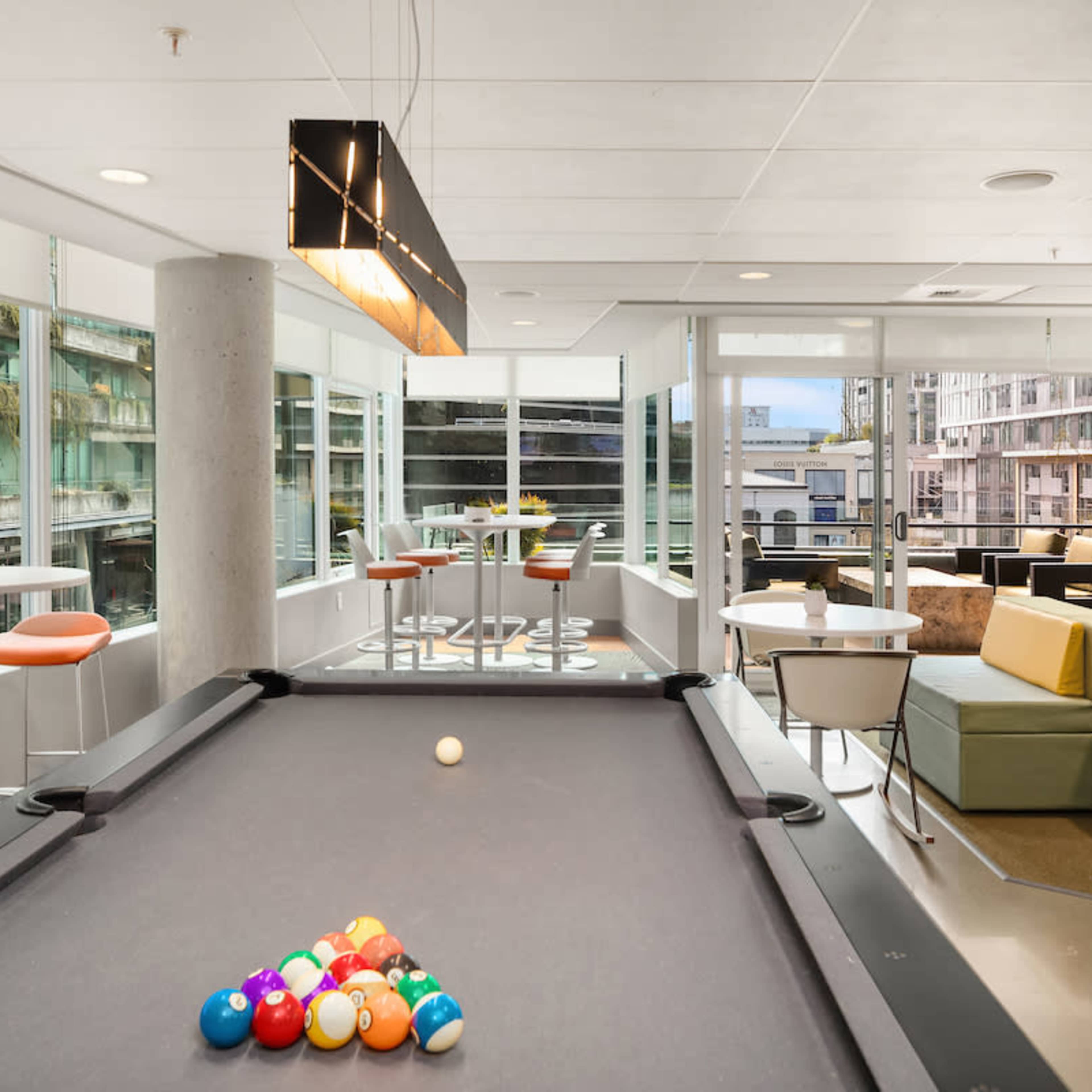 A modern game room featuring a billiards table with a set of colorful pool balls, surrounded by bright seating areas and large windows that allow natural light to enter.