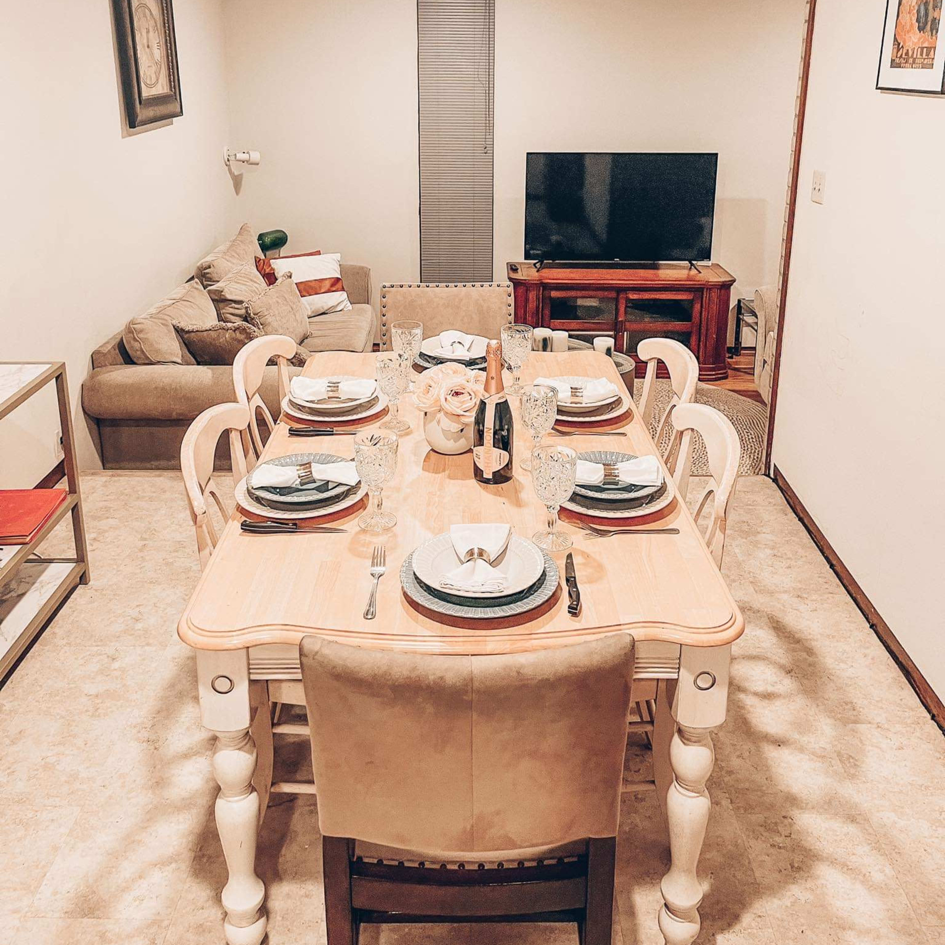 A dining area features a table set for eight with plates and glasses, a television, and a sofa in the background.